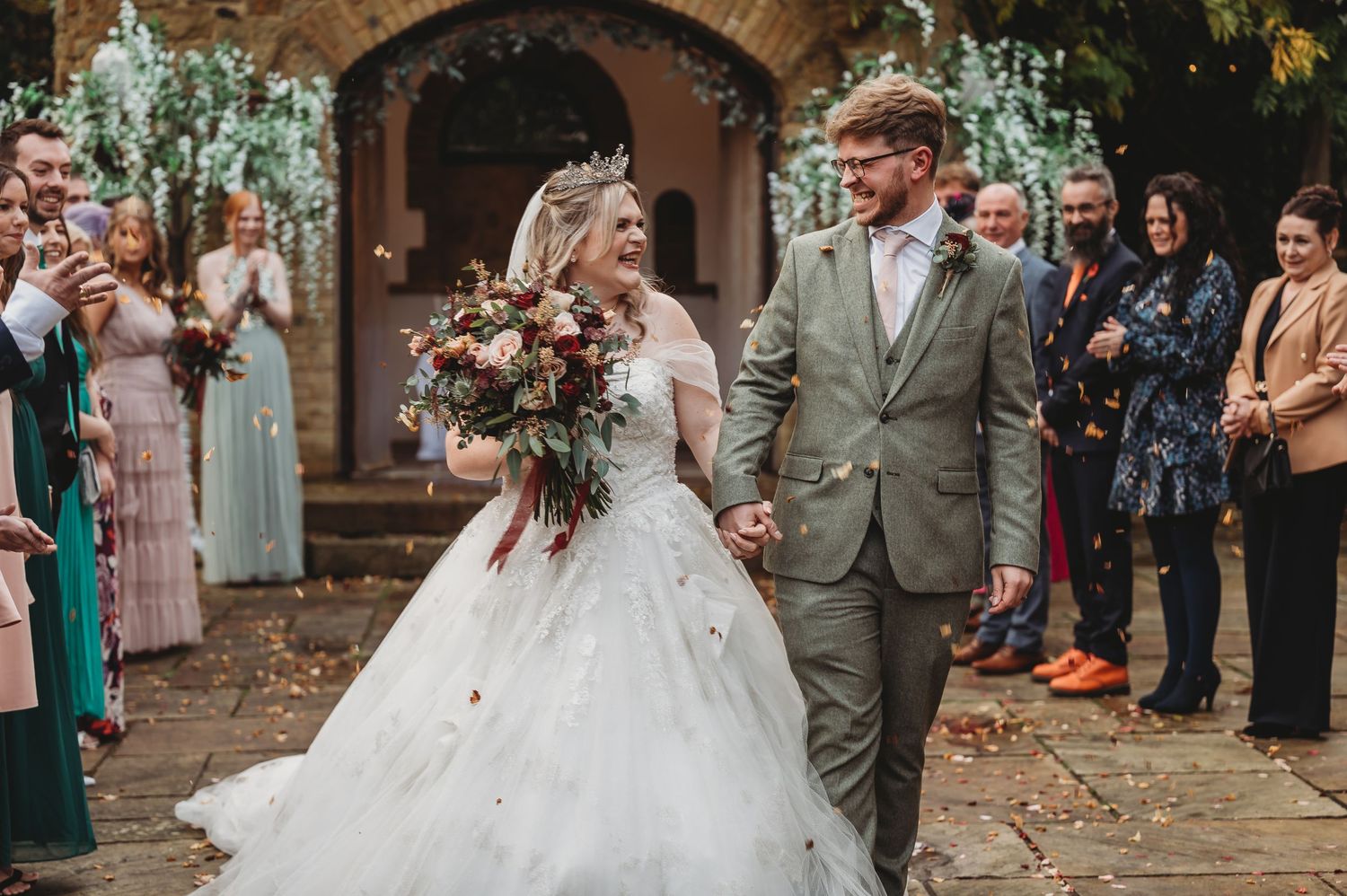 Wedding couple exits stone church as guests line pathway during autumn ceremony.
