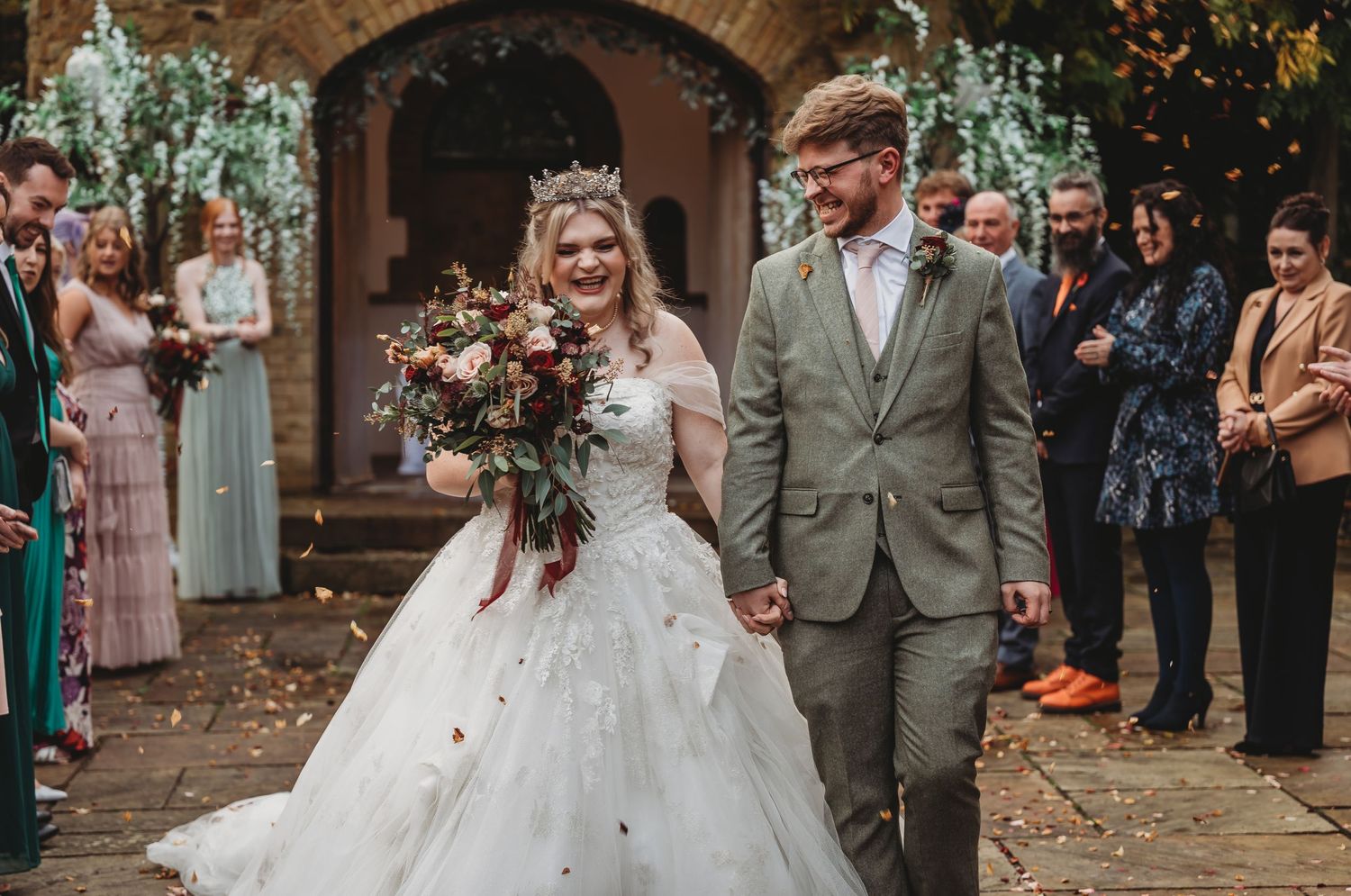 Newlyweds walk down church pathway while guests line sides during autumn wedding.