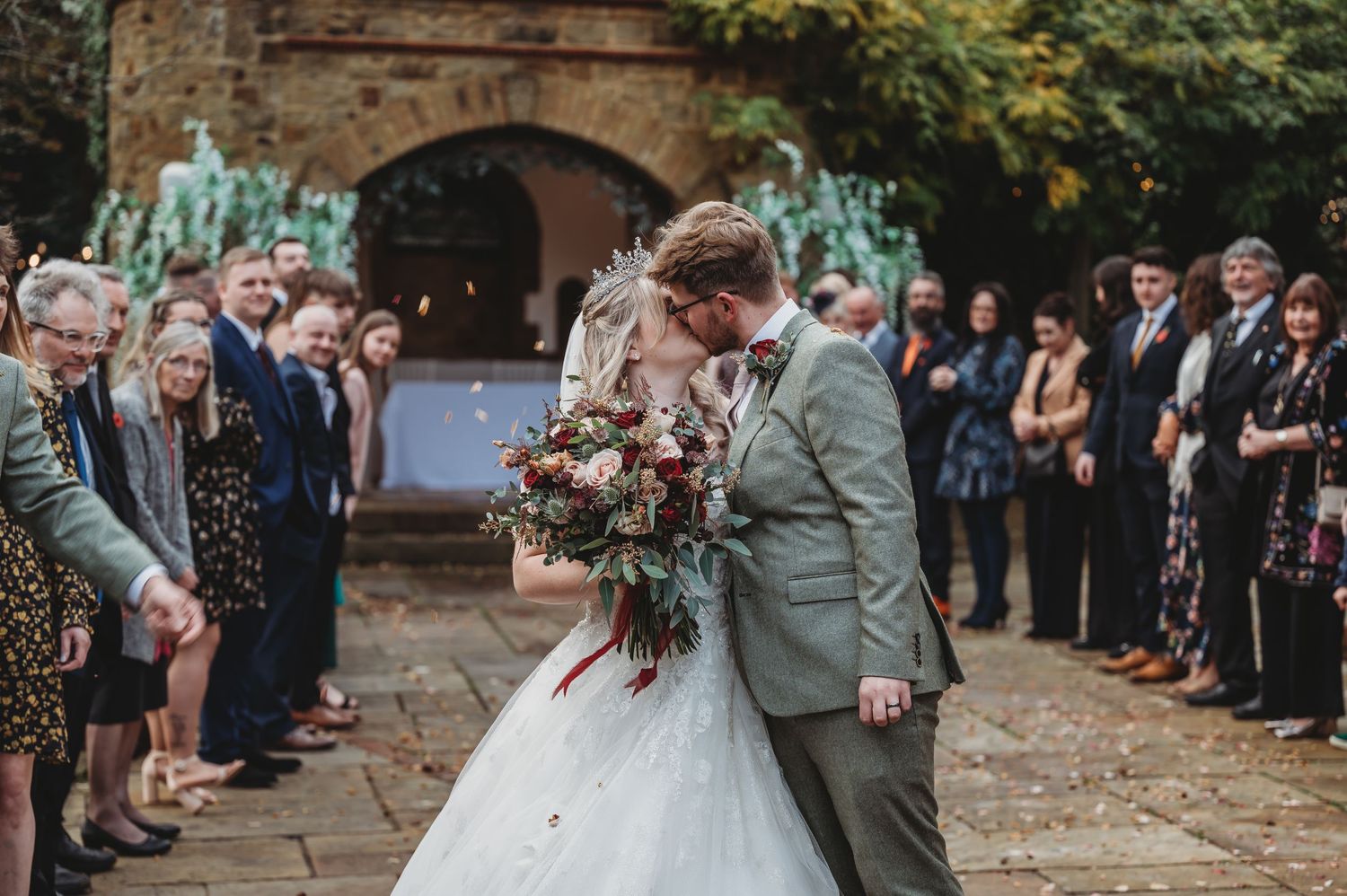 Wedding couple shares intimate moment as guests form aisle outside stone church.