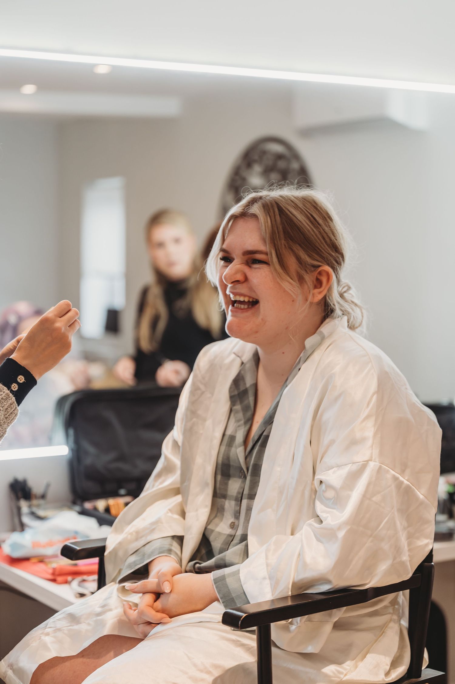 Someone laughing while sitting in a makeup chair wearing a white robe.