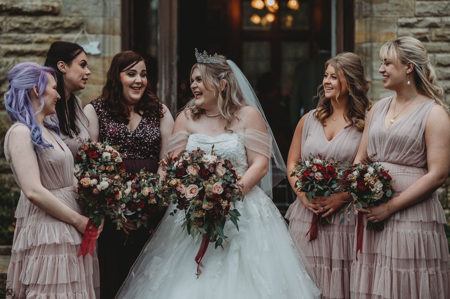 Wedding party in pink dresses shares laughter while holding beautiful floral bouquets.