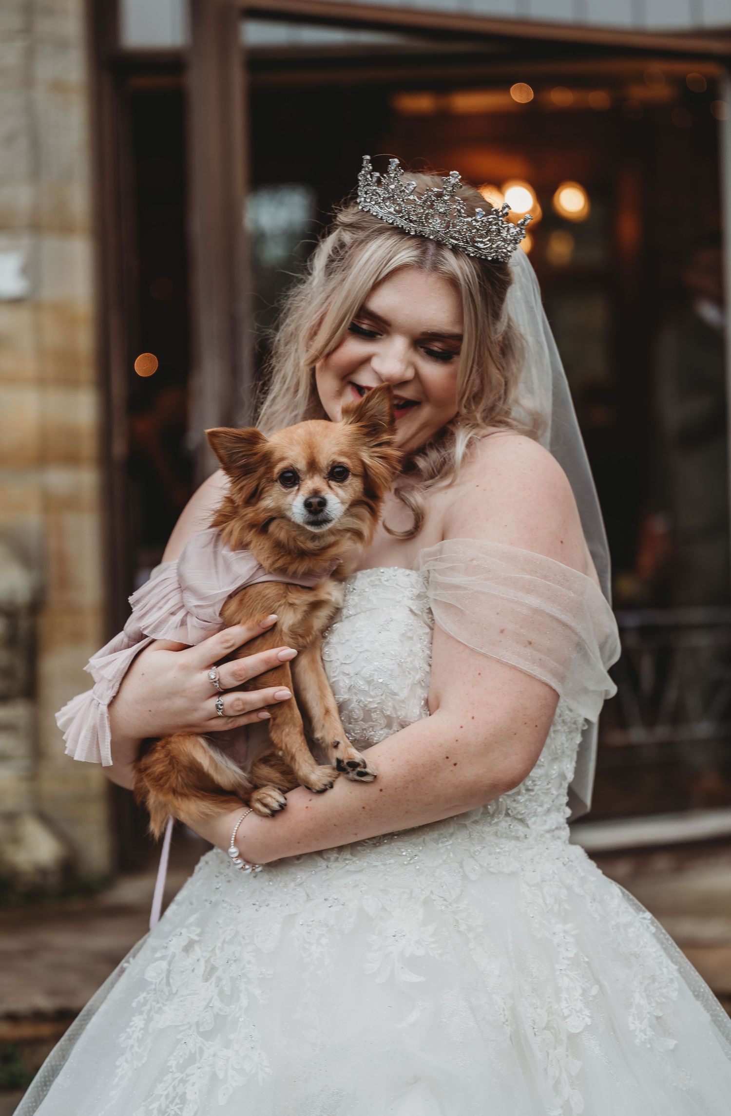 Joyful bride wearing a tiara and white gown holds her small Chihuahua dog during wedding photos.