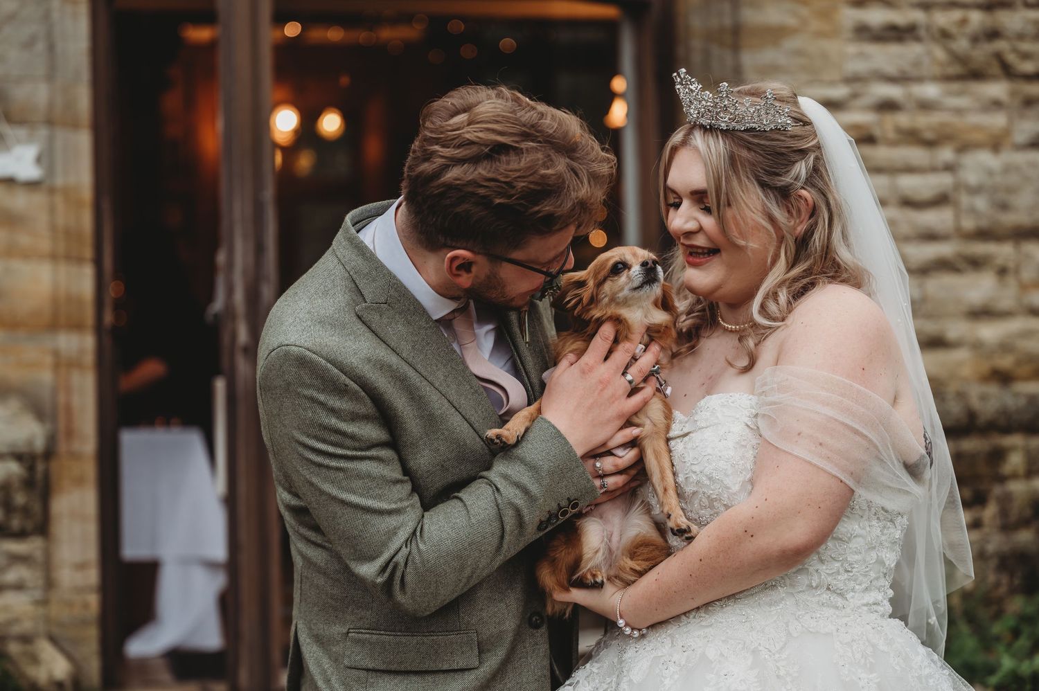 Happy couple cuddles their small dog during their wedding celebration by a stone wall.