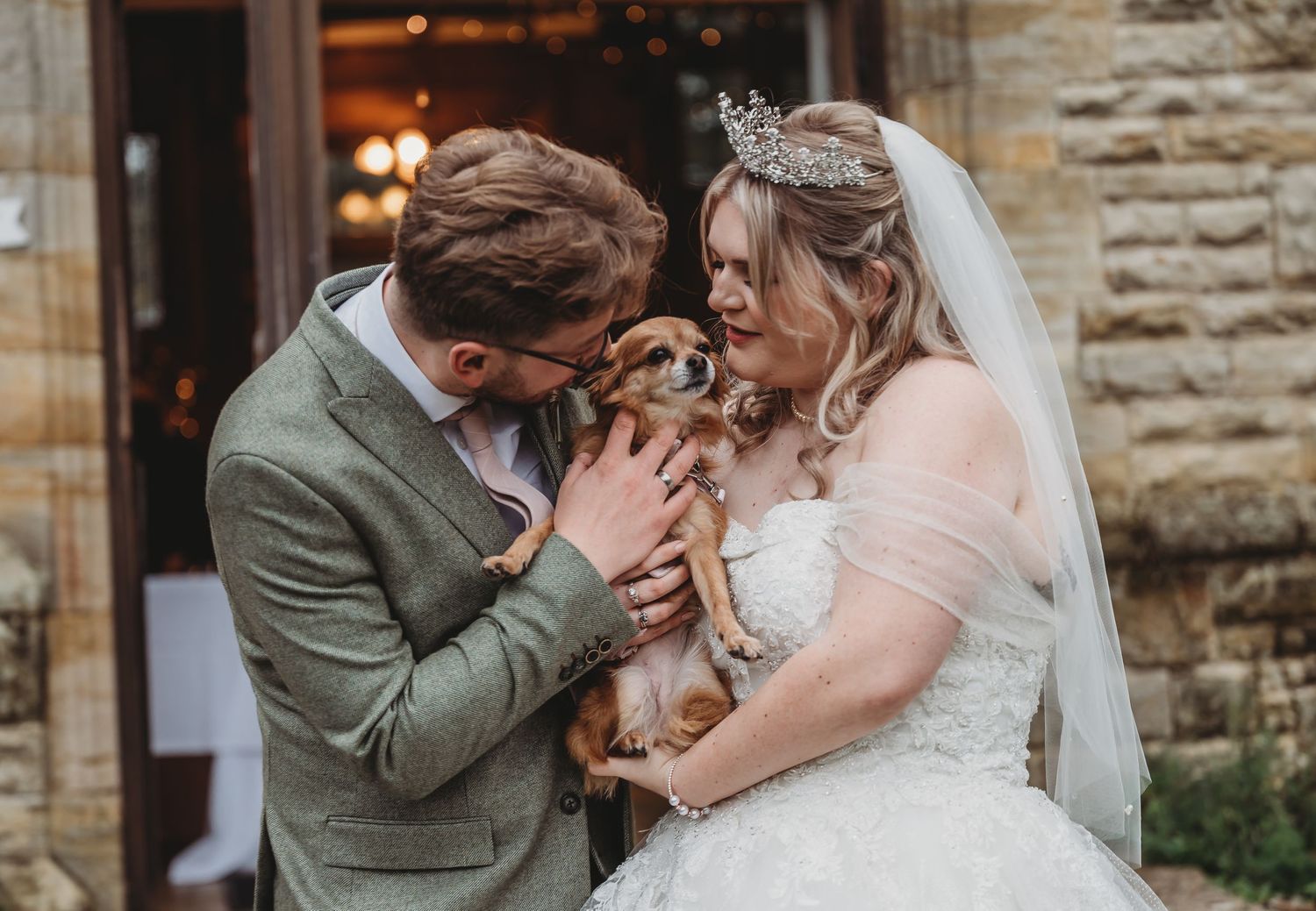 Newlywed couple in wedding attire share a joyful moment with their small dog outside a stone building.