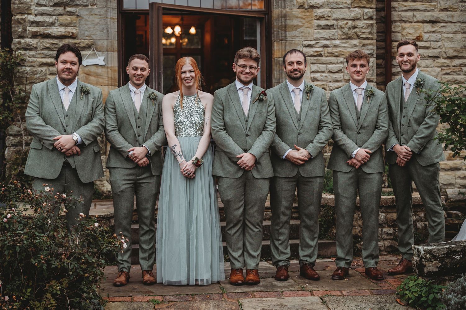 Wedding party in coordinated sage green attire poses formally on stone steps.