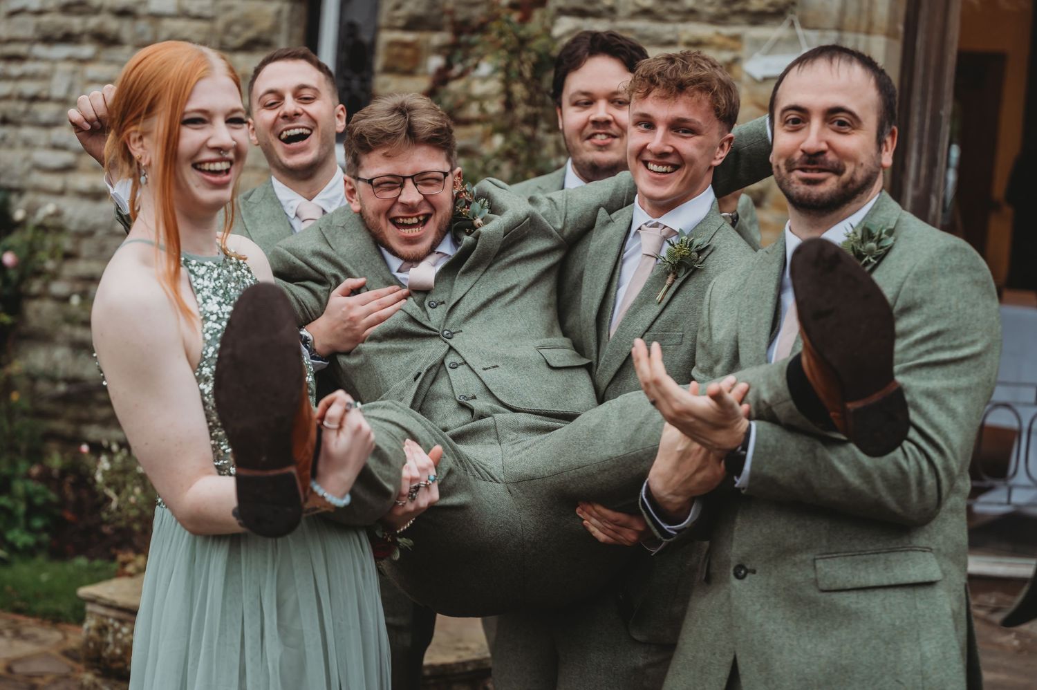 Wedding party in sage green attire share a playful moment lifting someone in celebration.