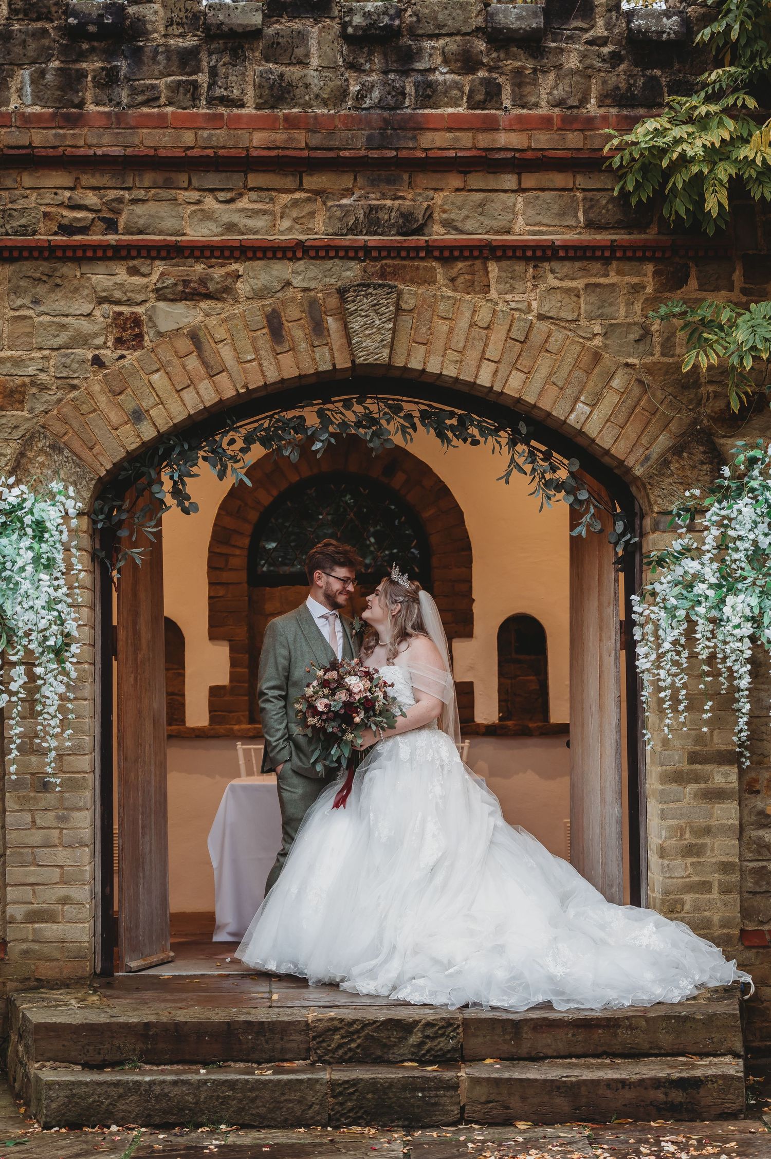 Elegant wedding portrait under a stone archway decorated with cascading white wisteria flowers.