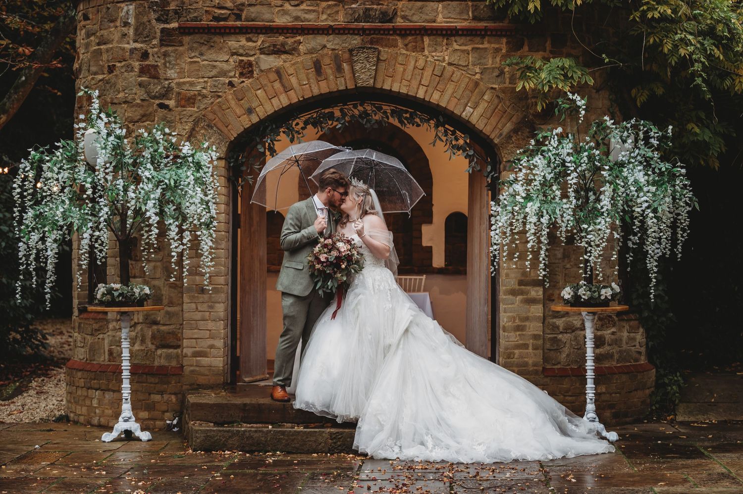 Romantic wedding photo featuring wisteria blooms framing an archway with a couple sharing an umbrella.