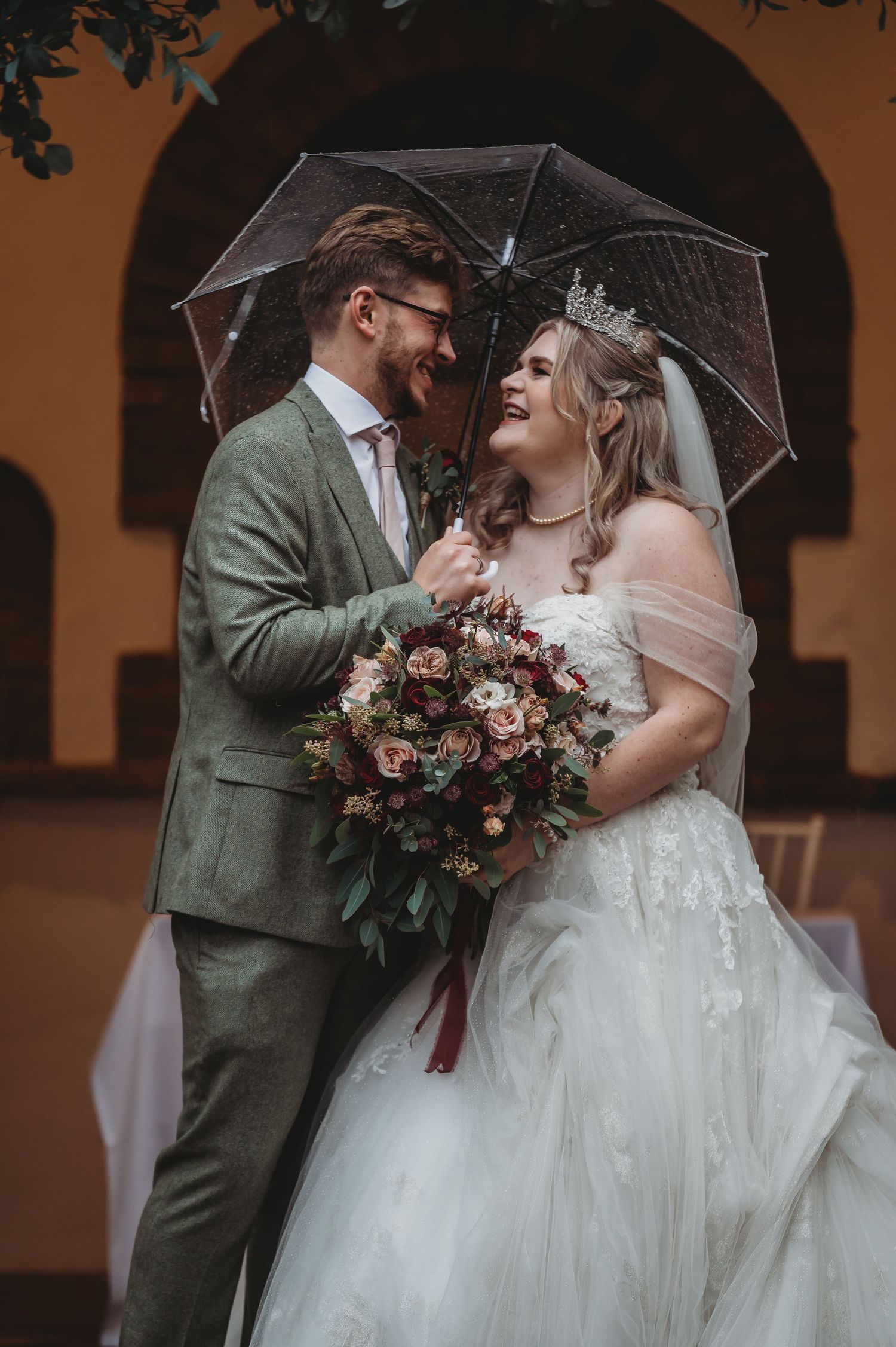 Wedding couple shares an intimate moment under a clear umbrella with a lush burgundy bouquet.