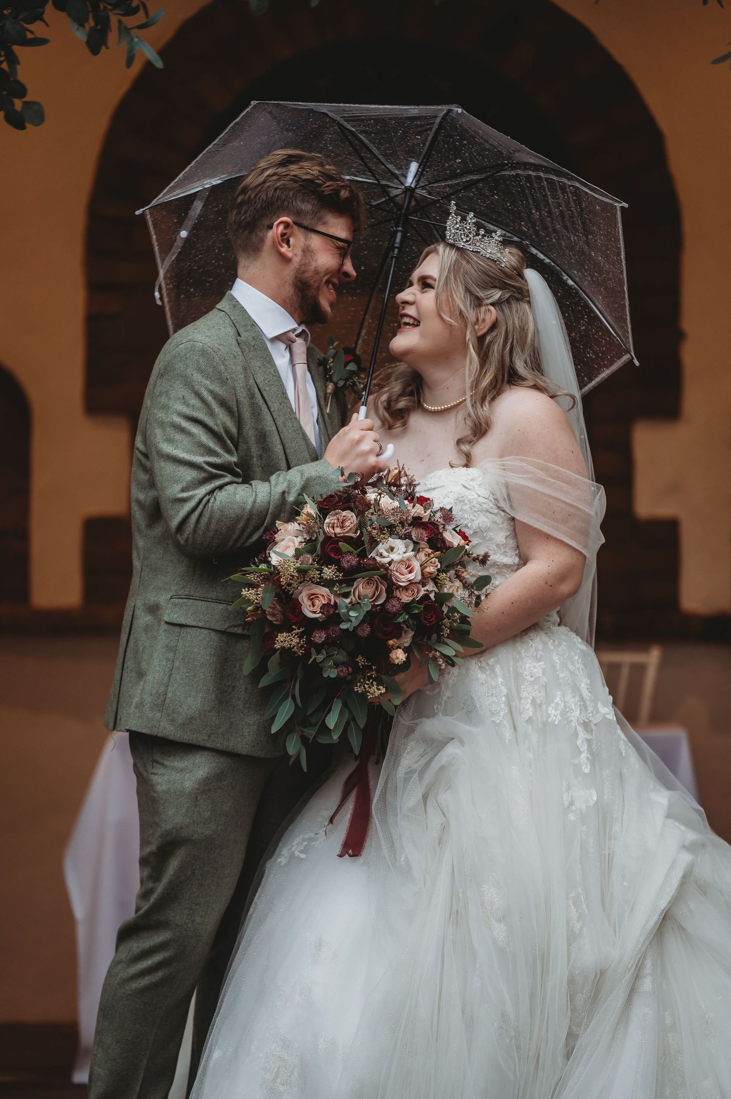 A joyful wedding moment shared under a clear umbrella against an arched architectural backdrop.