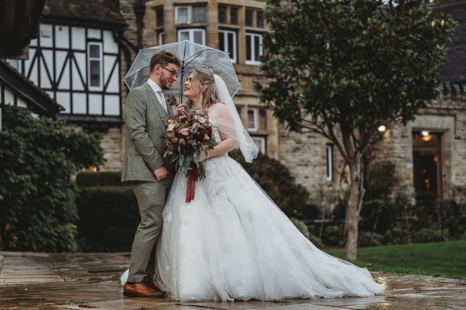 Elegant wedding couple pose together against stone architecture while holding a burgundy floral bouquet.