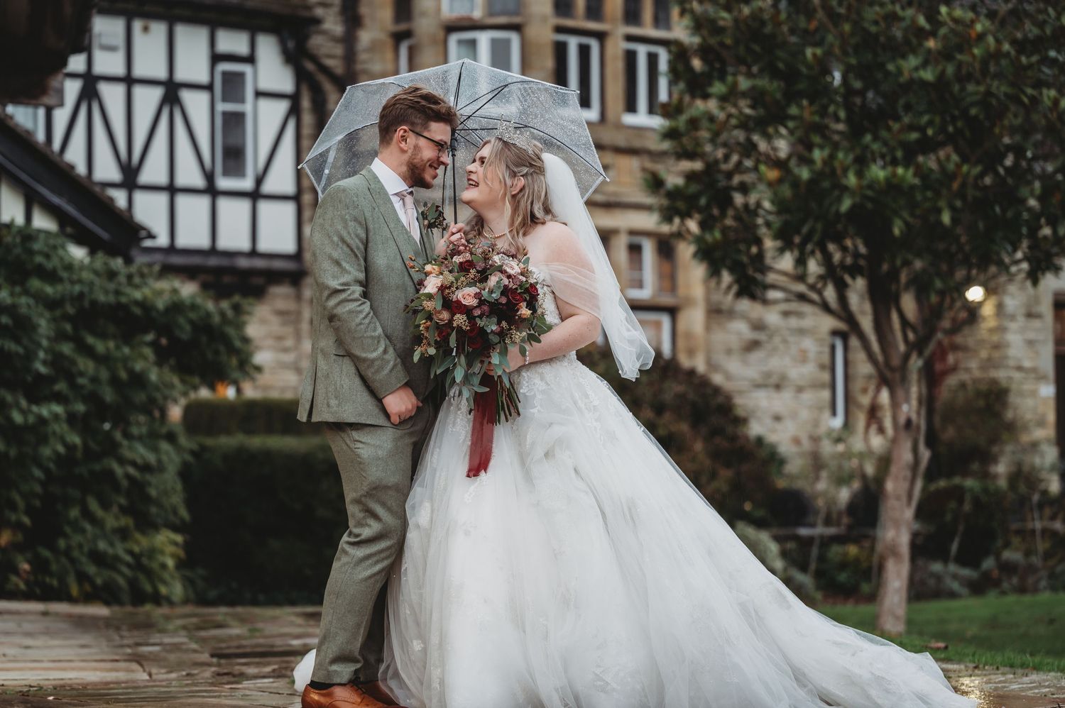 A romantic wedding portrait in front of a tudor-style building featuring a flowing white gown and sage green suit.