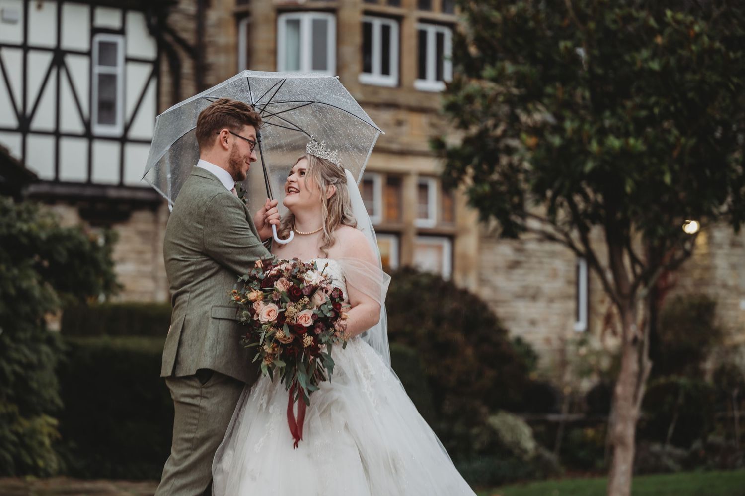 Couple poses with umbrella and bouquet outside historic Tudor building on rainy day.