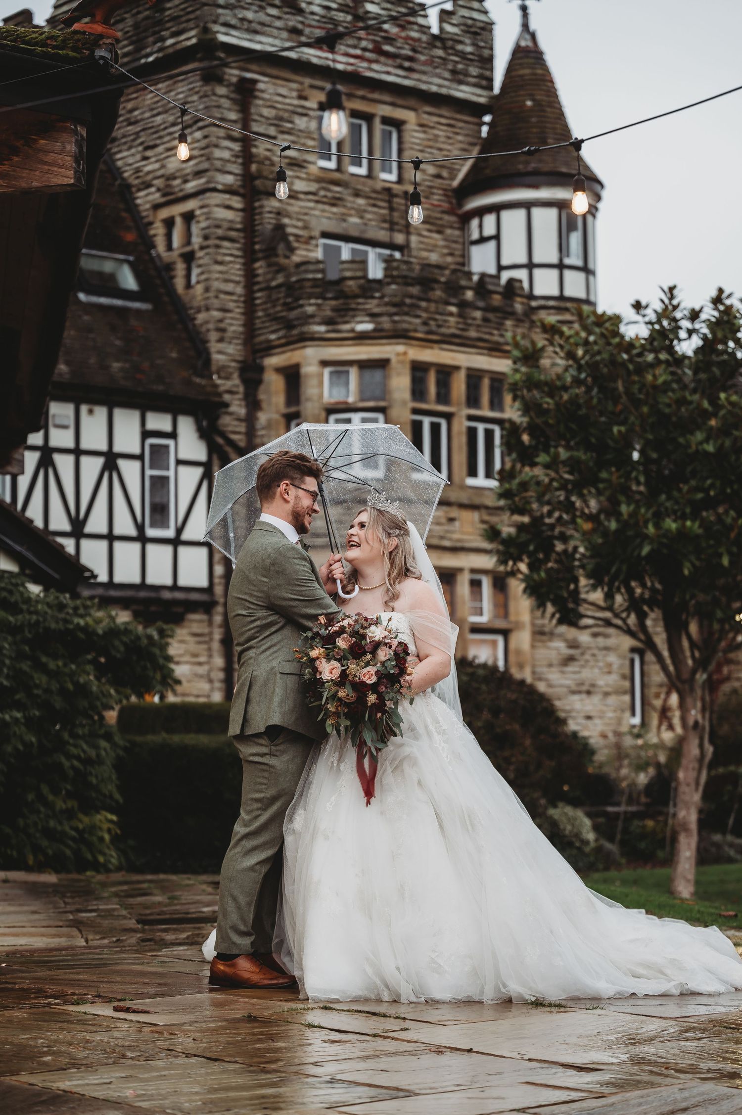 Couple shares romantic moment under clear umbrella outside stone Tudor-style mansion.