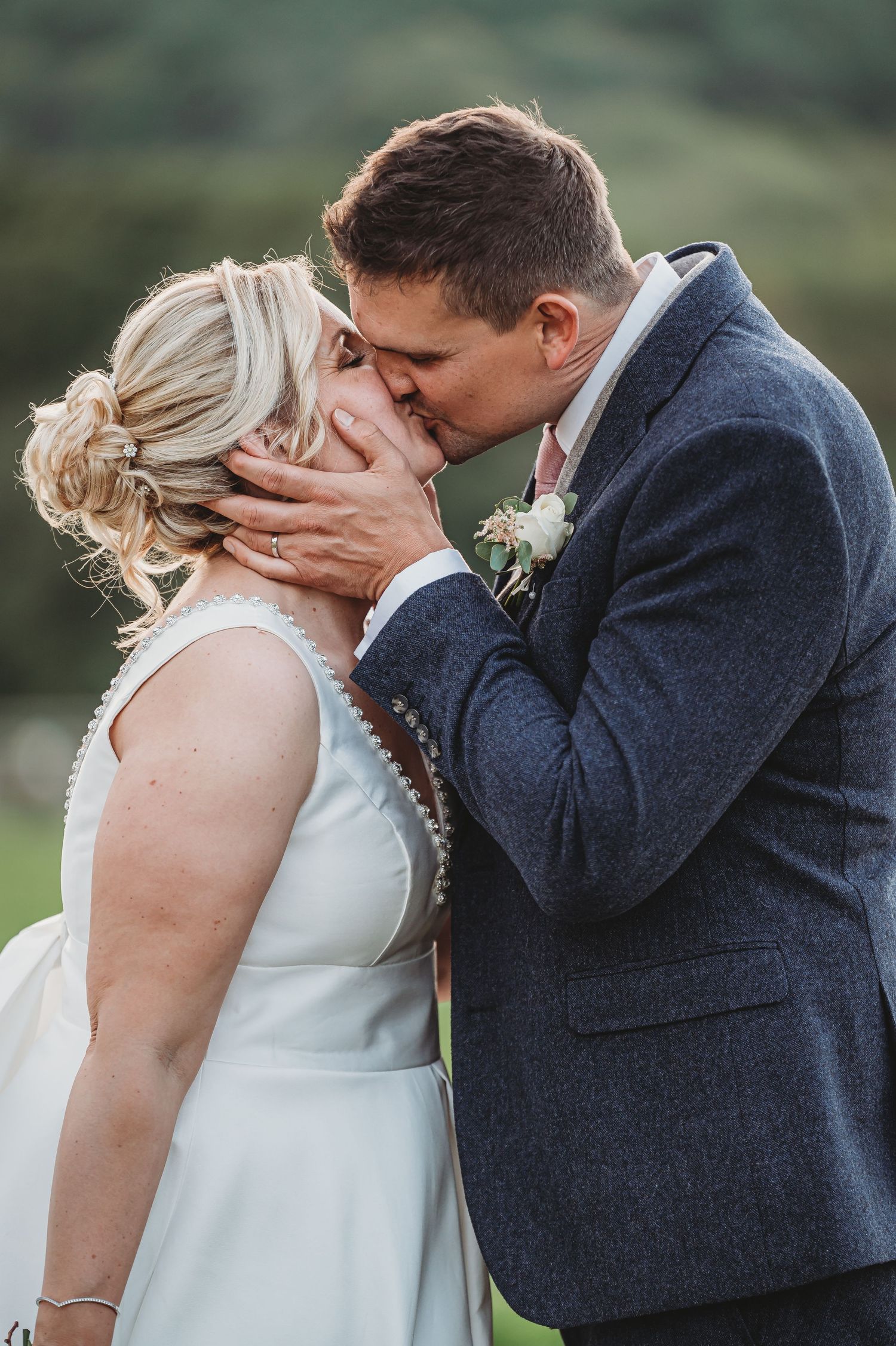 A romantic wedding couple share a passionate kiss outdoors, with the bride in white and groom in a navy suit.