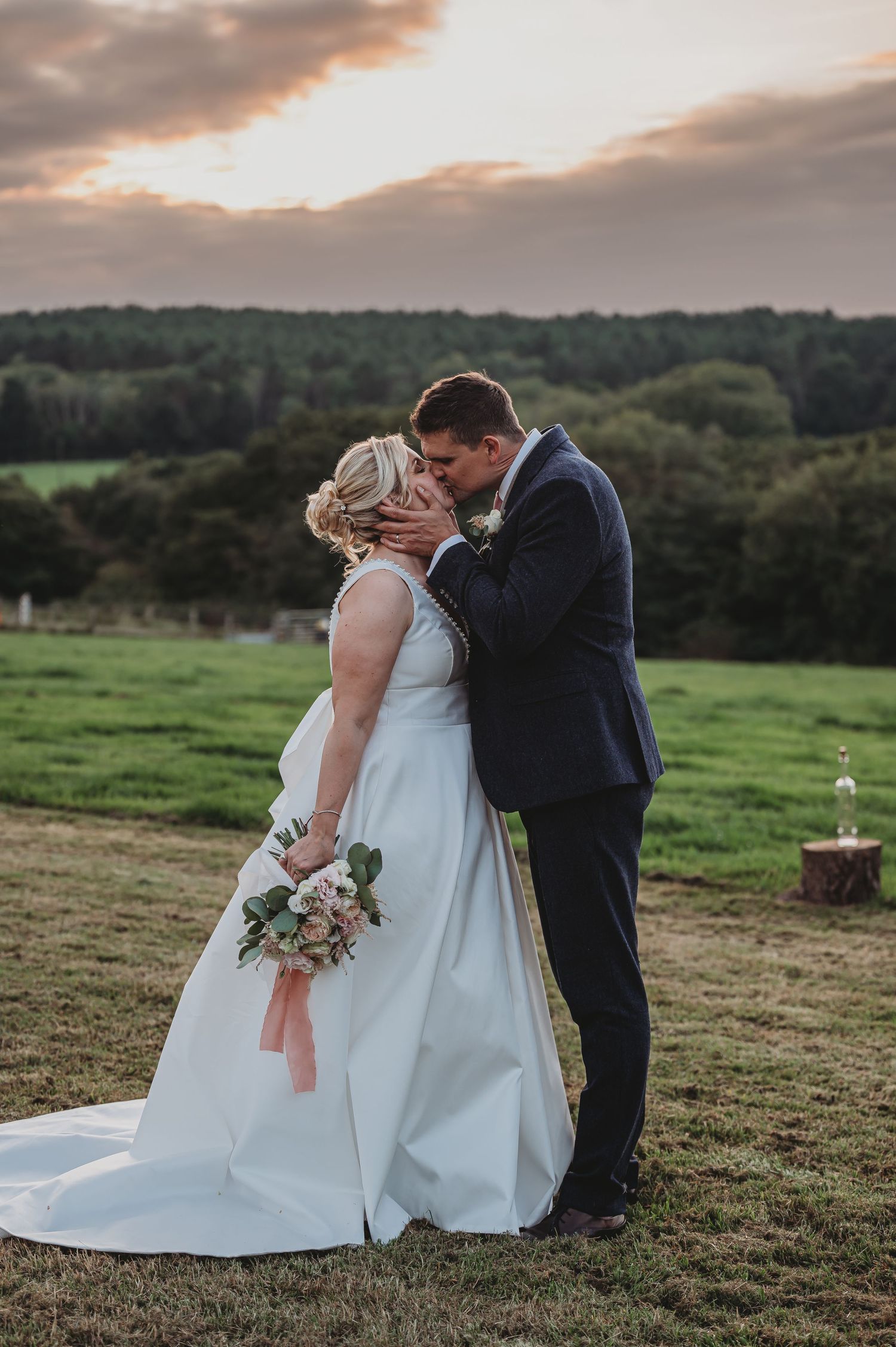 A romantic sunset wedding photo showing a couple embracing on a hilltop overlooking lush green fields and trees.