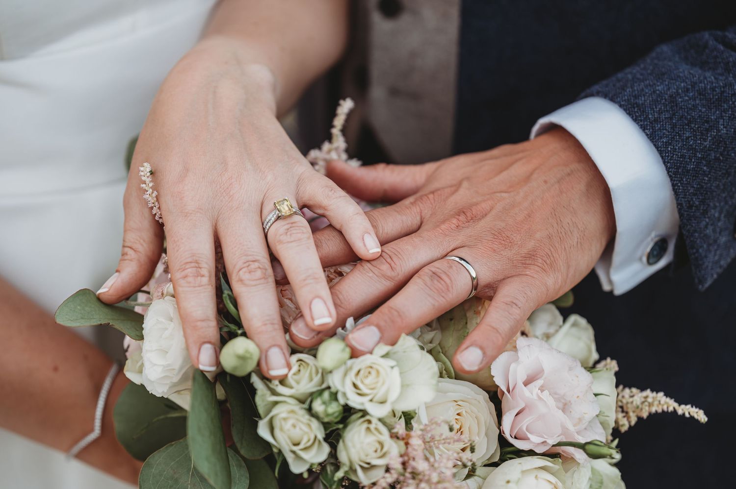 Newlywed couple's hands with wedding rings resting on a bouquet of white and pink roses.