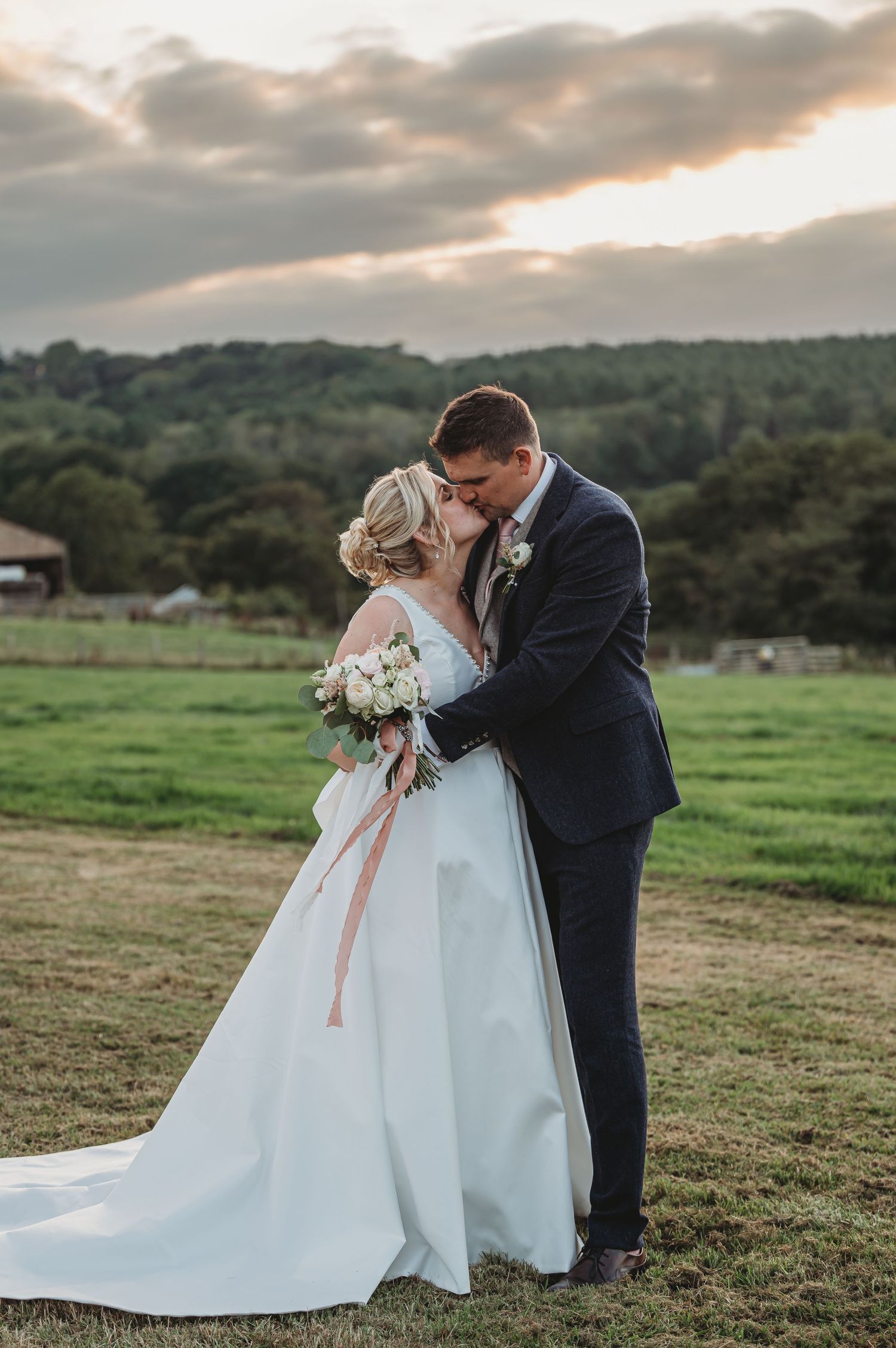 A romantic wedding couple embraces in a secluded meadow during golden hour with rolling hills and soft clouds in the background.