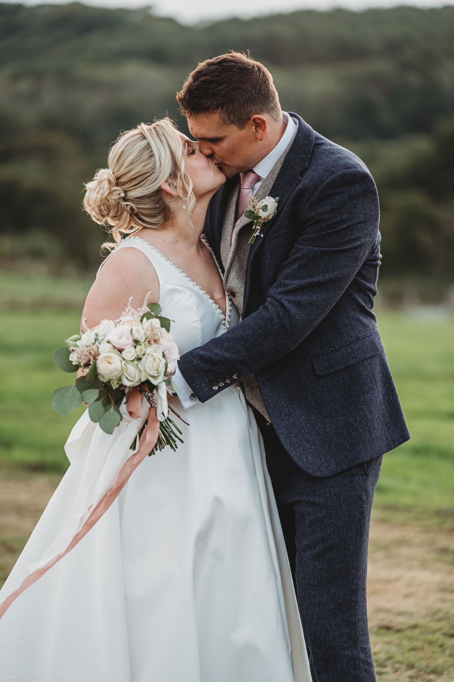 A romantic wedding moment outdoors with the bride in white holding a blush pink bouquet and groom in navy suit.