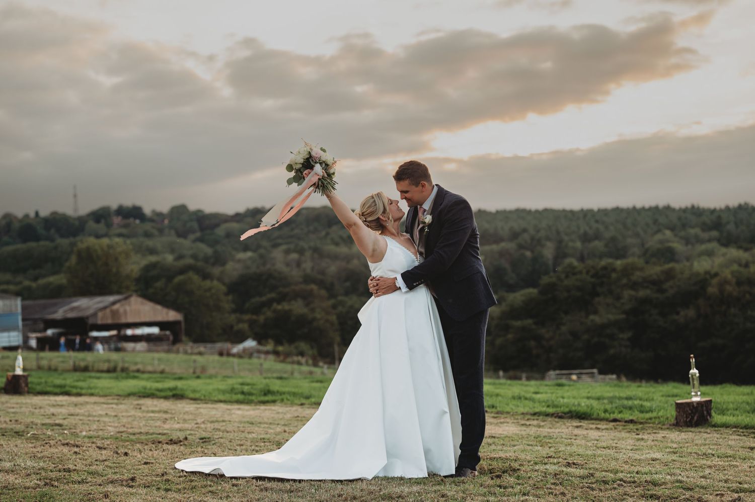 A couple in wedding attire embrace on a hilltop meadow during sunset with trees and cloudy skies in the background.