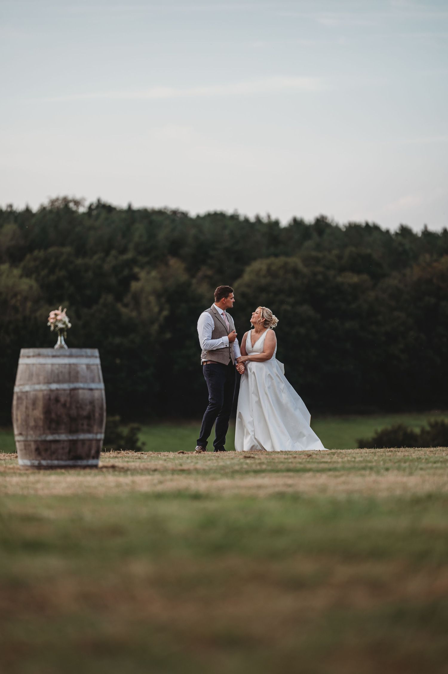 Rustic outdoor wedding scene with couple standing next to wine barrel in field with forest backdrop at sunset.