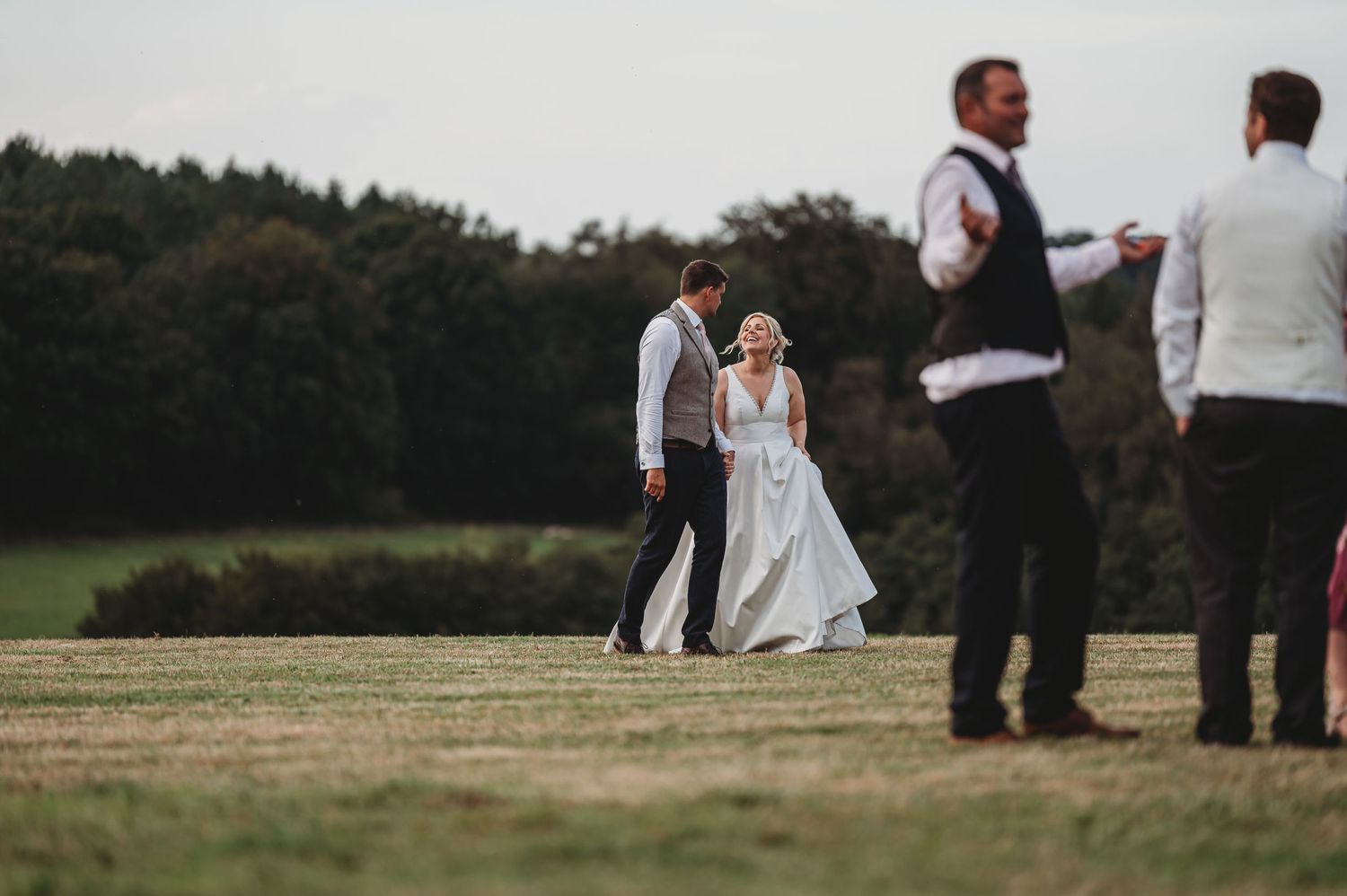 Wedding party members gather in a grassy field with trees in the background during outdoor reception celebration.