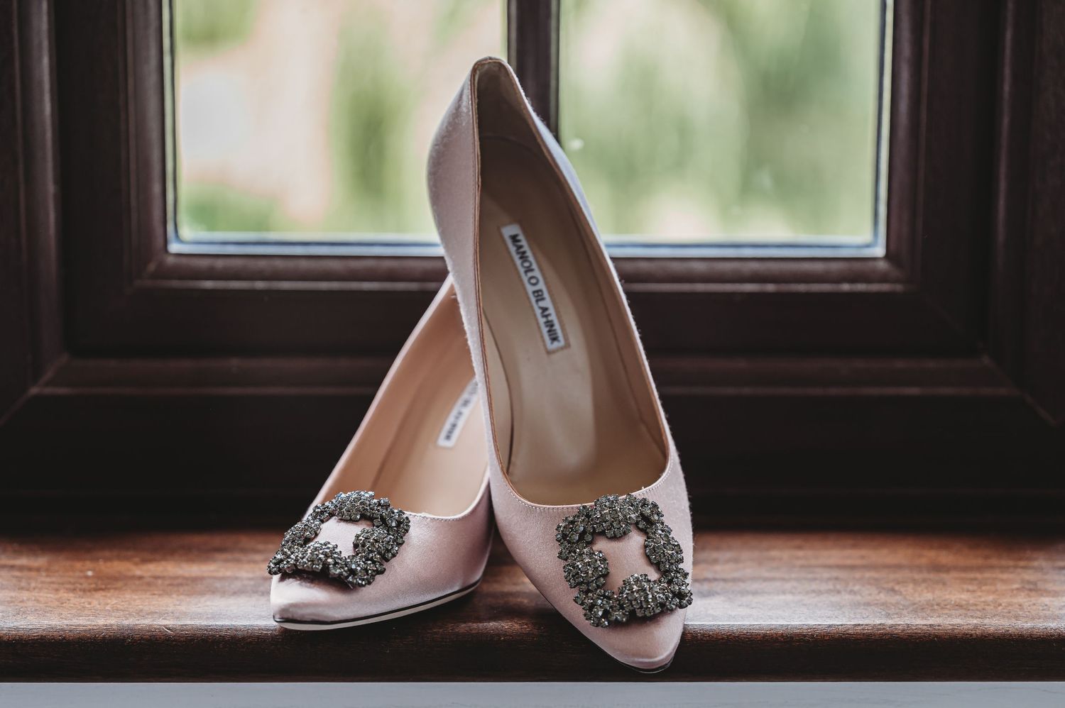 Elegant pink satin wedding shoes with ornate crystal brooches sitting on wooden windowsill in natural light.