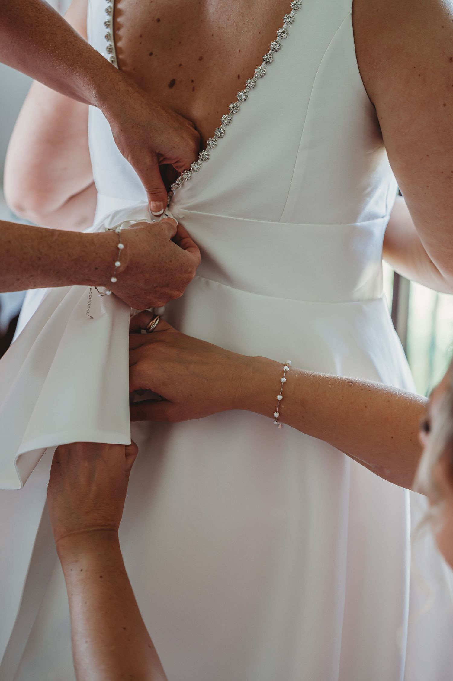 Hands adjusting and tying up the back of an elegant white wedding dress with delicate lace trim and pearl bracelets.