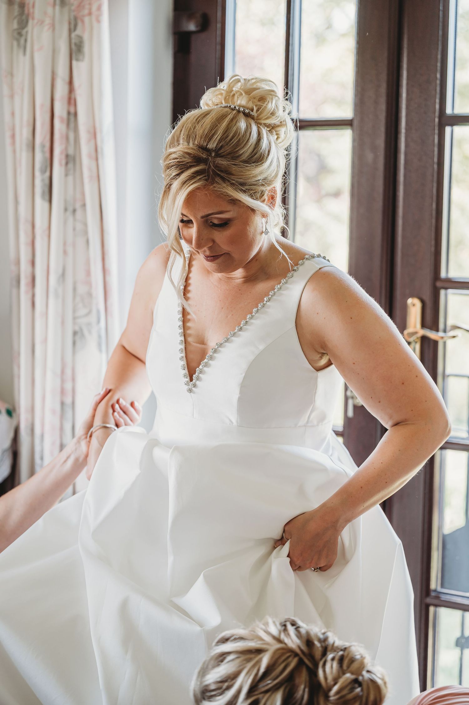 A bride in an elegant white V-neck wedding dress adjusts her gown while getting ready by a window with natural light.