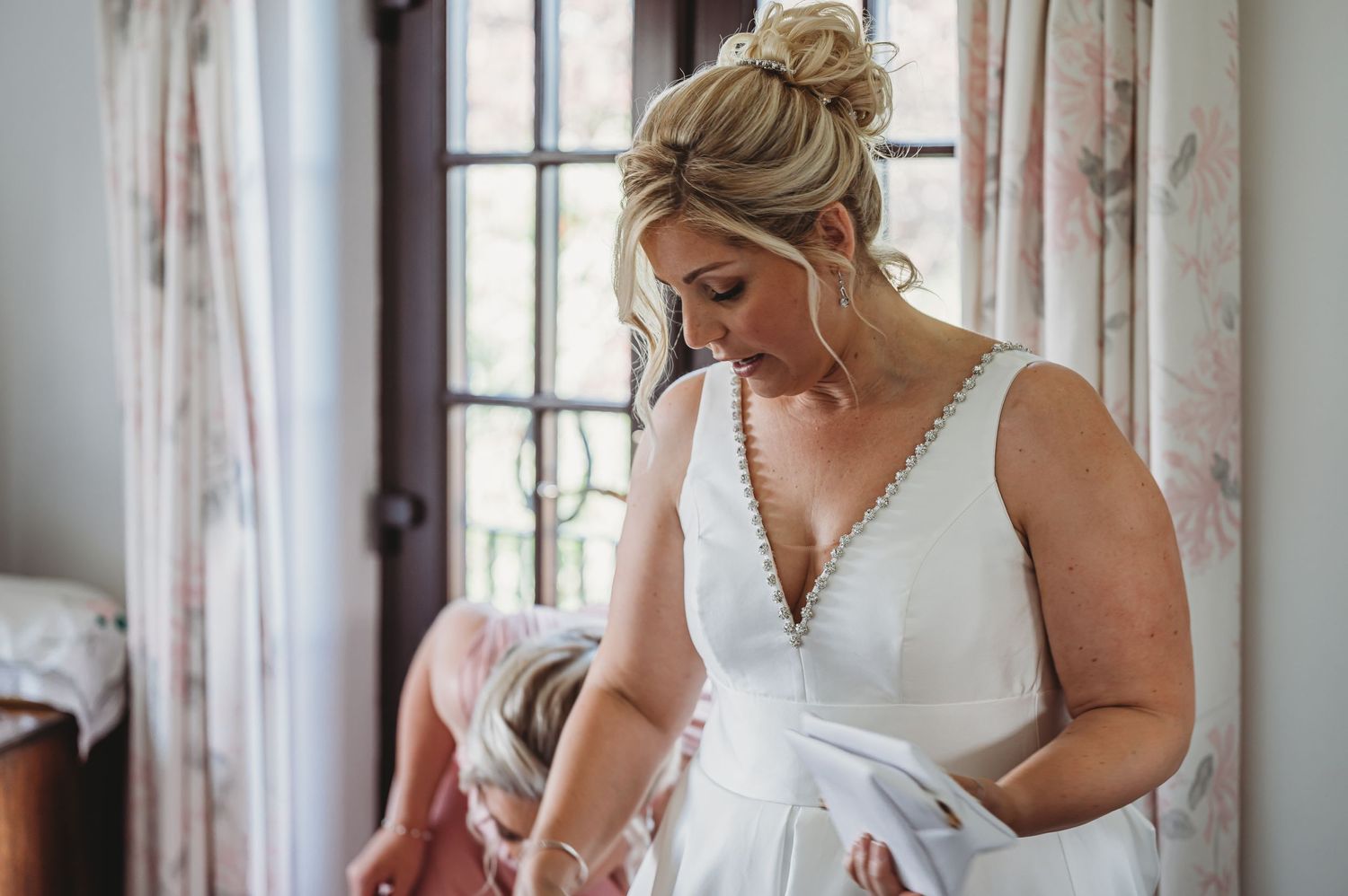 Someone getting ready before a wedding ceremony in a white bridal gown with deep v-neck detail by a window.