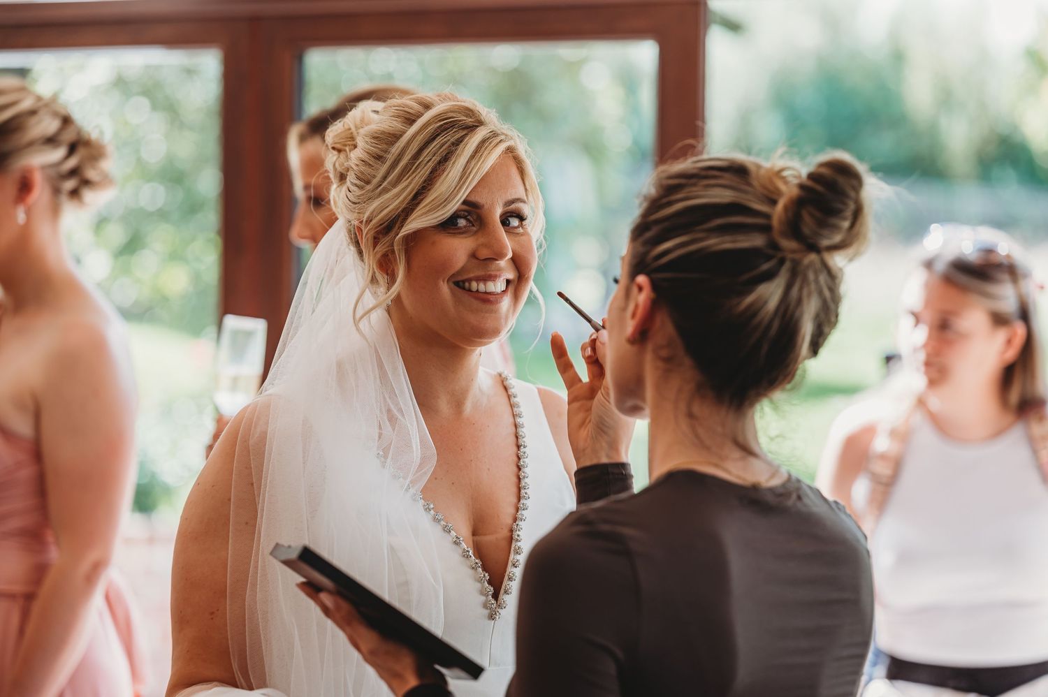 A makeup artist applies finishing touches to a bride wearing a white dress and veil during wedding preparations.