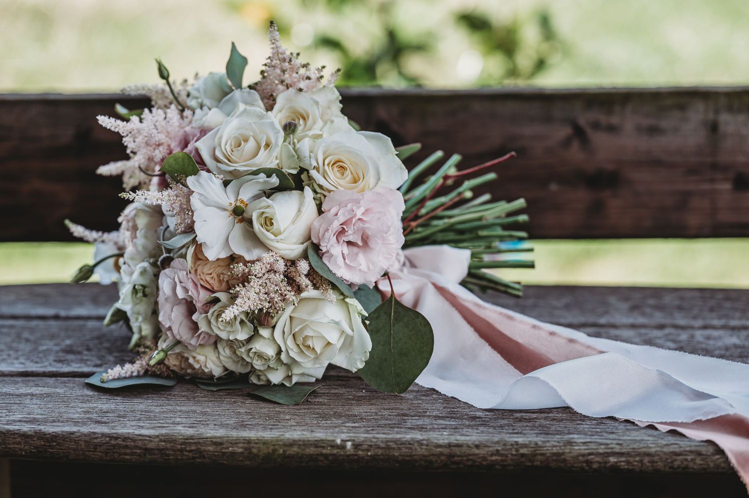 A delicate bridal bouquet with white roses, pink accents, and silvery greenery tied with a blush ribbon rests on wooden bench.