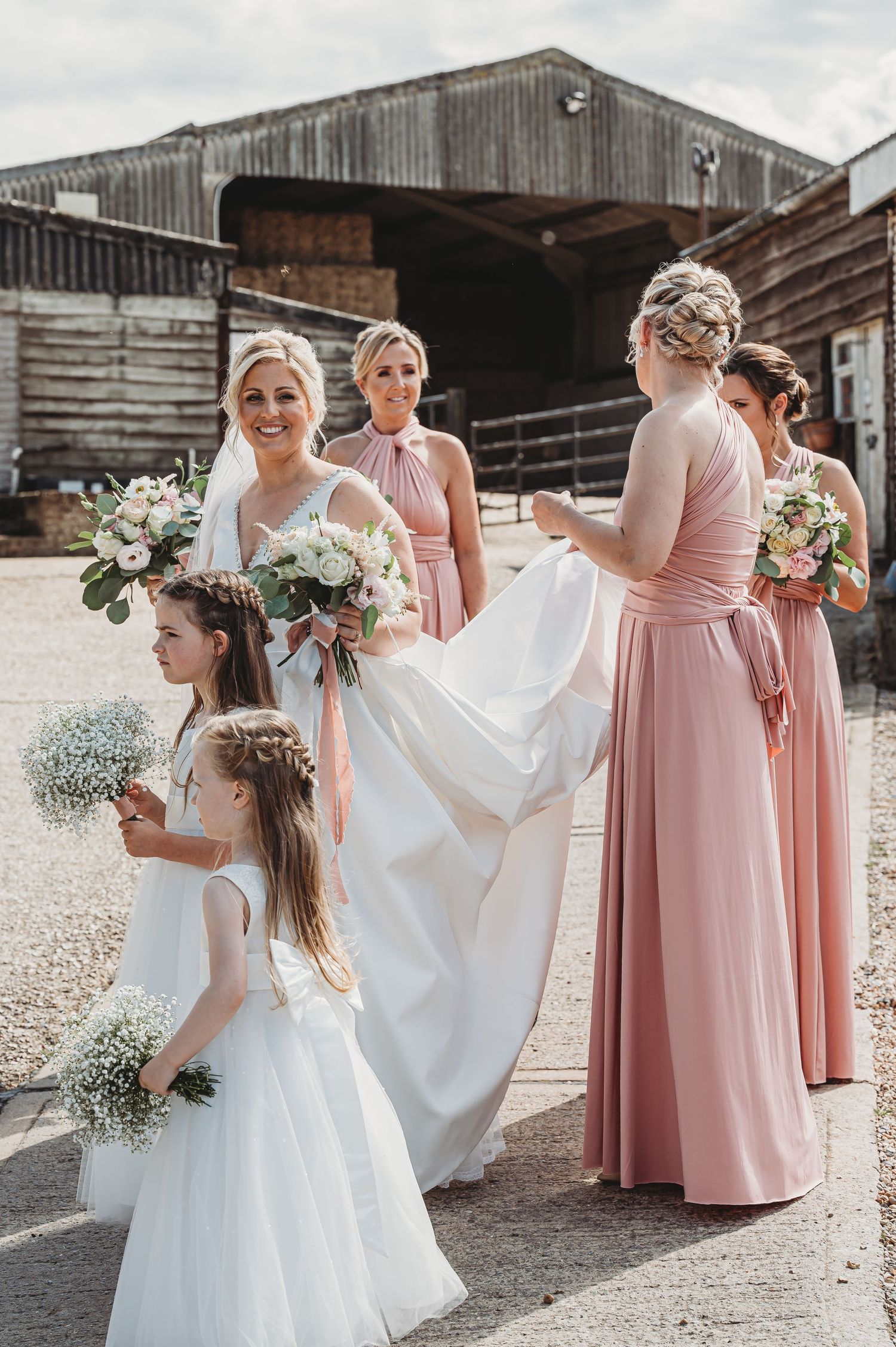 Wedding party wearing pink bridesmaid dresses and white gowns gather outside rustic barn venue with floral bouquets.