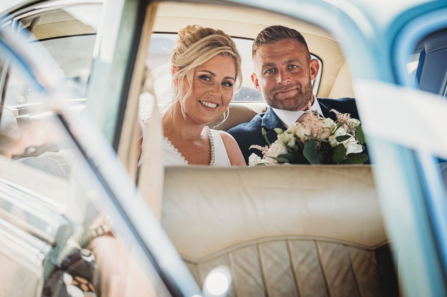 Newlyweds smile together in back seat of vintage car with white floral bouquet.