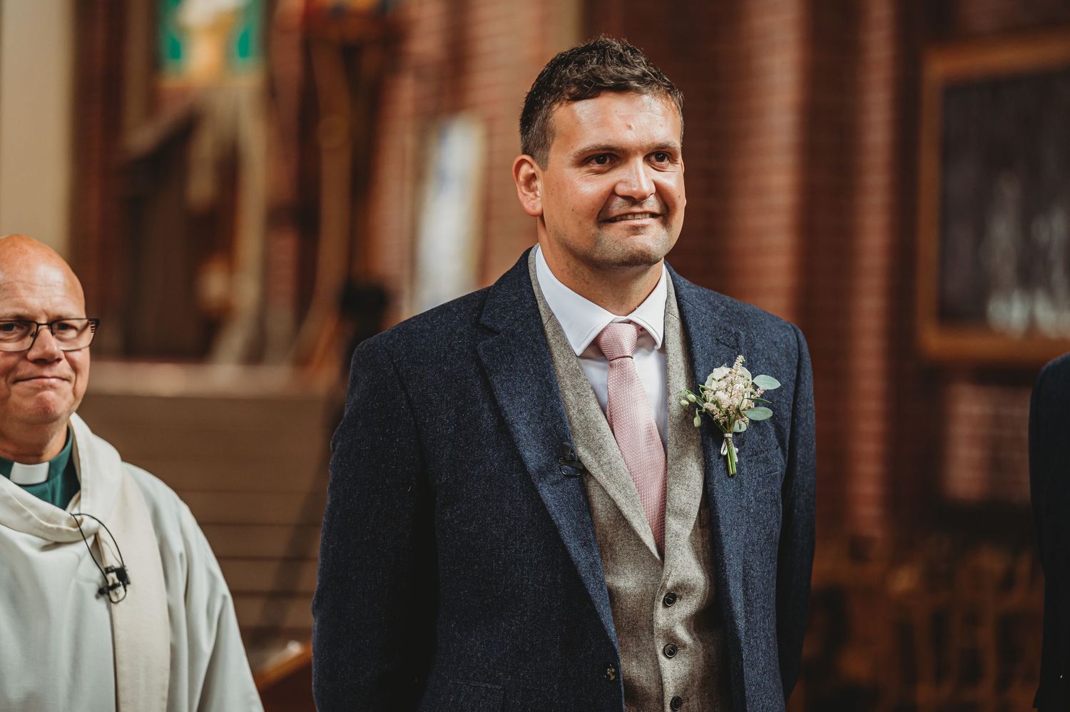 A groom wearing a navy suit with pink tie stands at the entrance of a brick church.