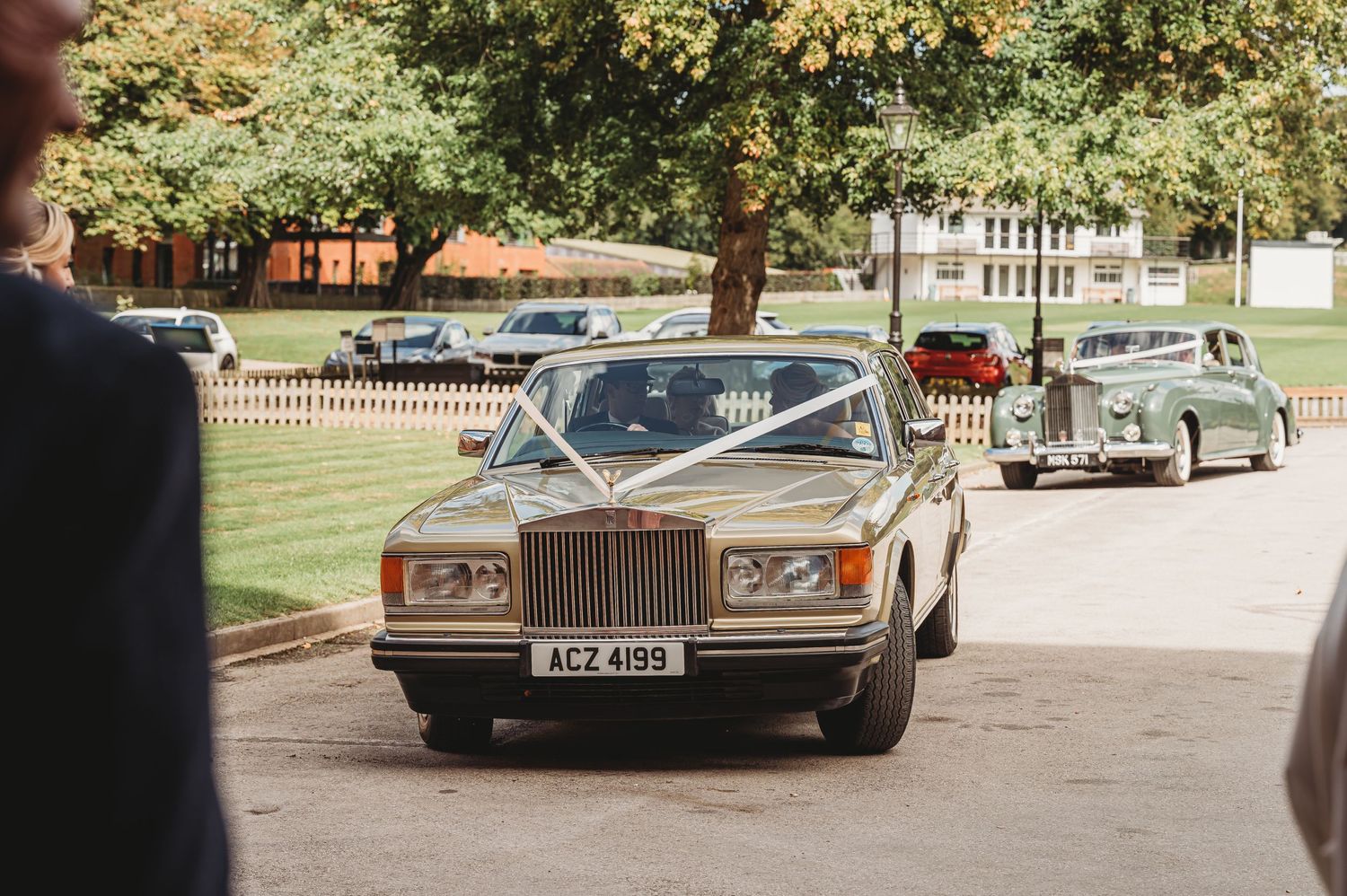A vintage Rolls-Royce Silver Spirit wedding car leading a classic vehicle procession on a tree-lined street.