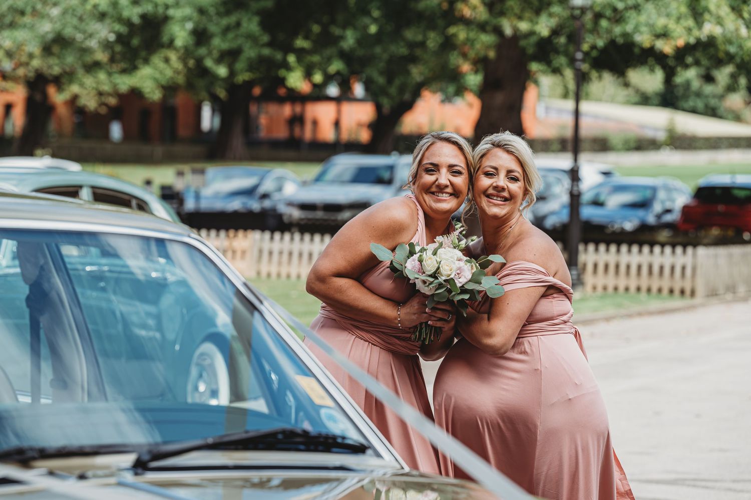 Two bridesmaids in pink dresses sharing a joyful moment outside with a bouquet.