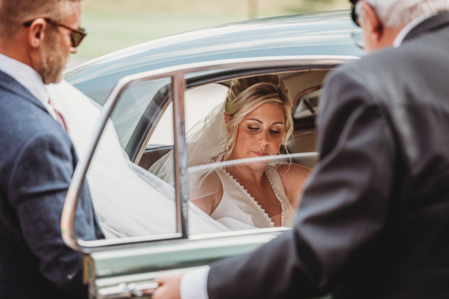 Bride in white wedding dress sitting inside vintage car viewed through window on wedding day.