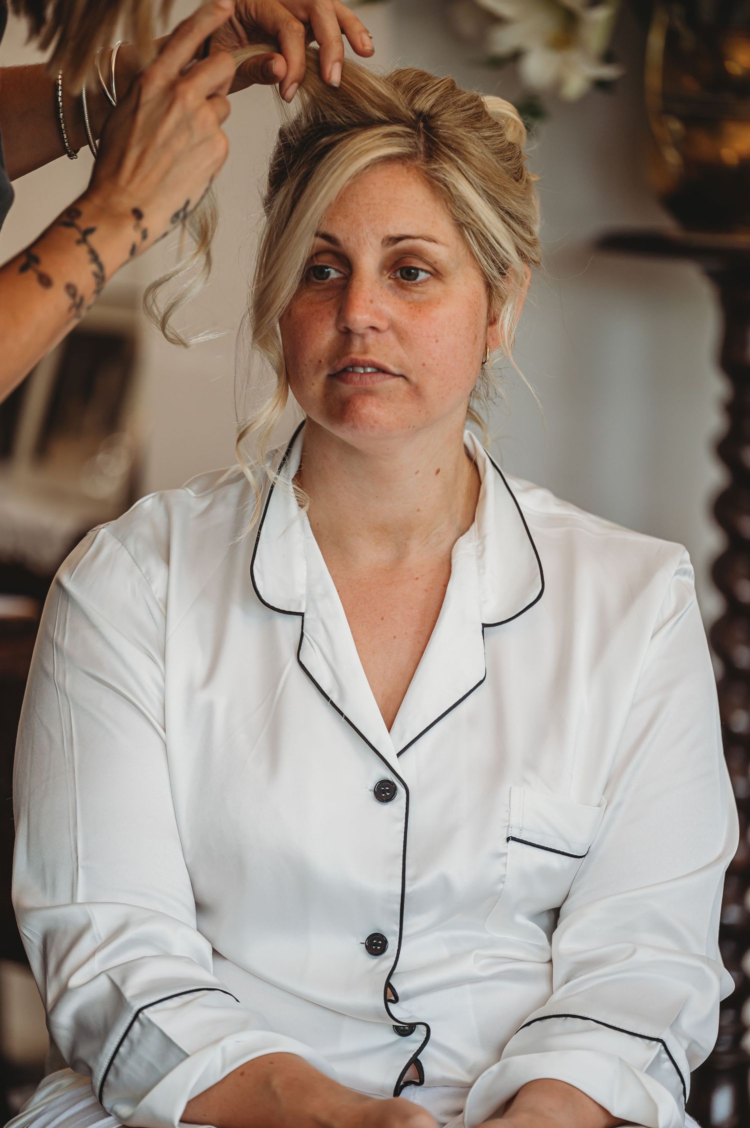 A person wearing a white button-up pajama top sits while having their hair styled by someone's hands.