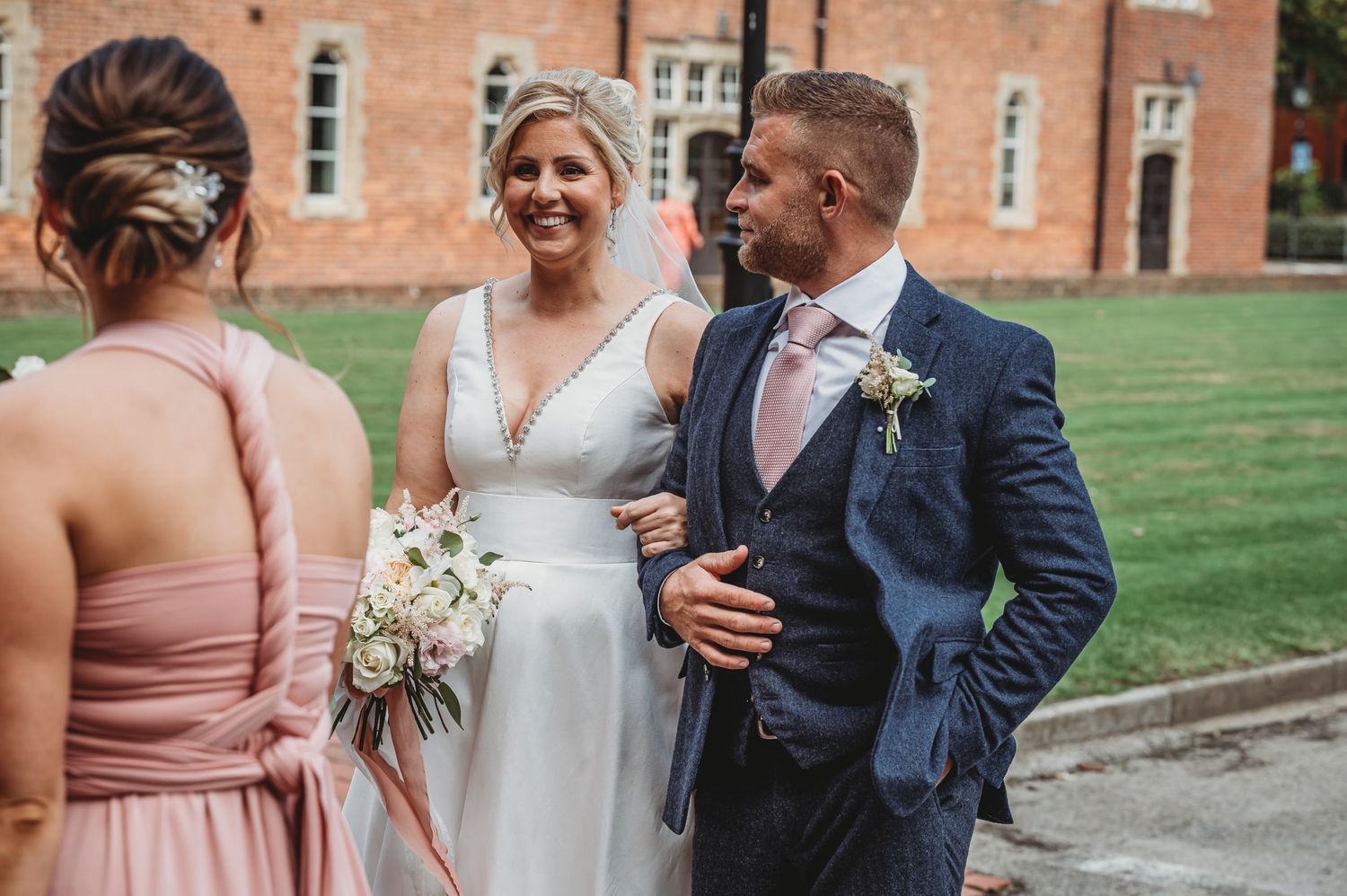 A joyful wedding scene with bridal party members in formal attire standing on green lawn outside brick building.