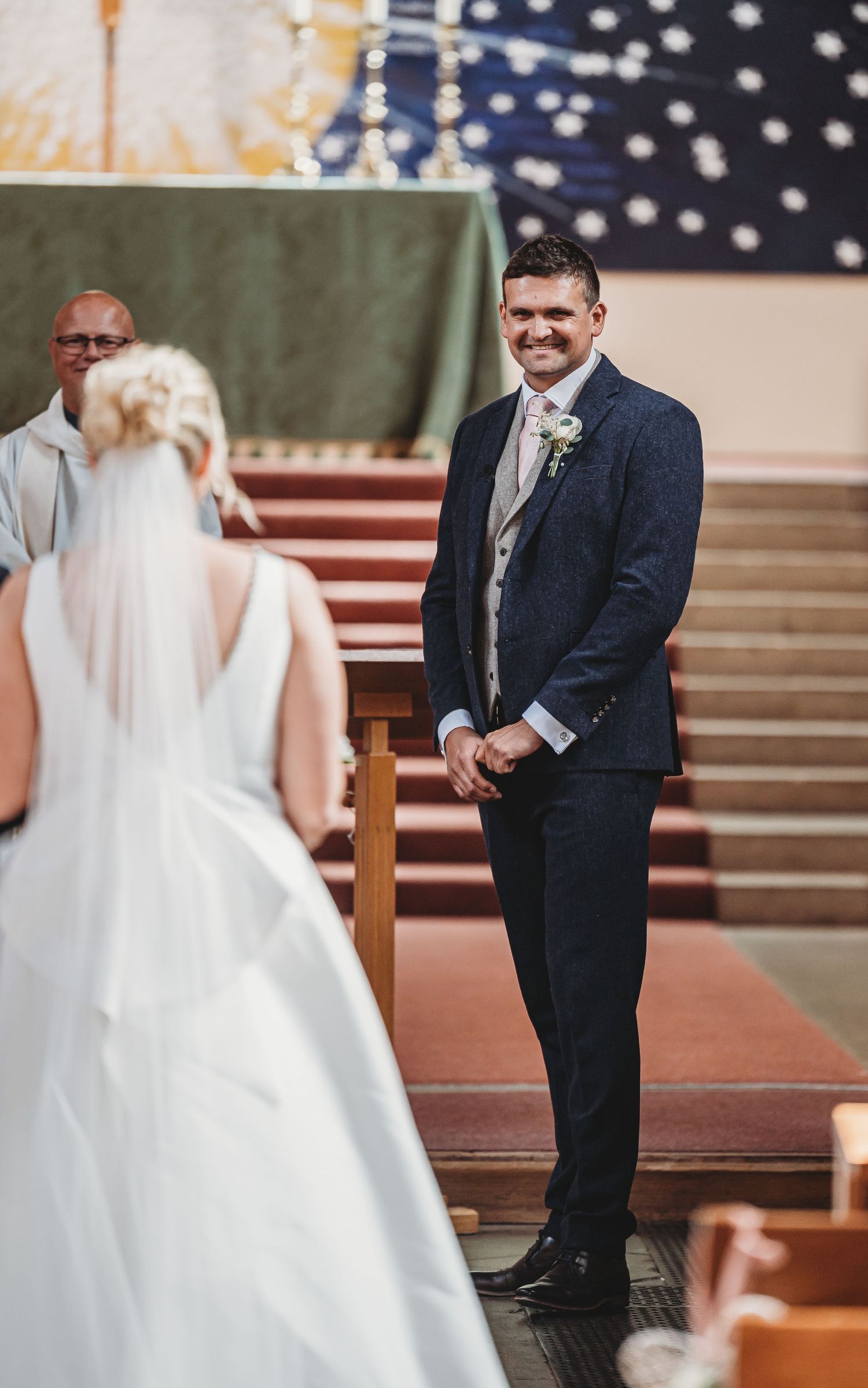 A wedding ceremony in a church with a groom in a navy suit standing at the altar looking at his bride in a white gown.