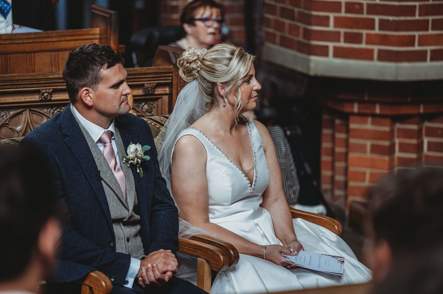 A couple sits together in wooden chairs inside a brick-walled church during a ceremony.