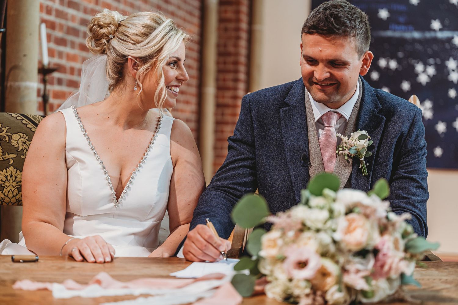 A couple signs documents at a table with a bridal bouquet of pink and white flowers on display.