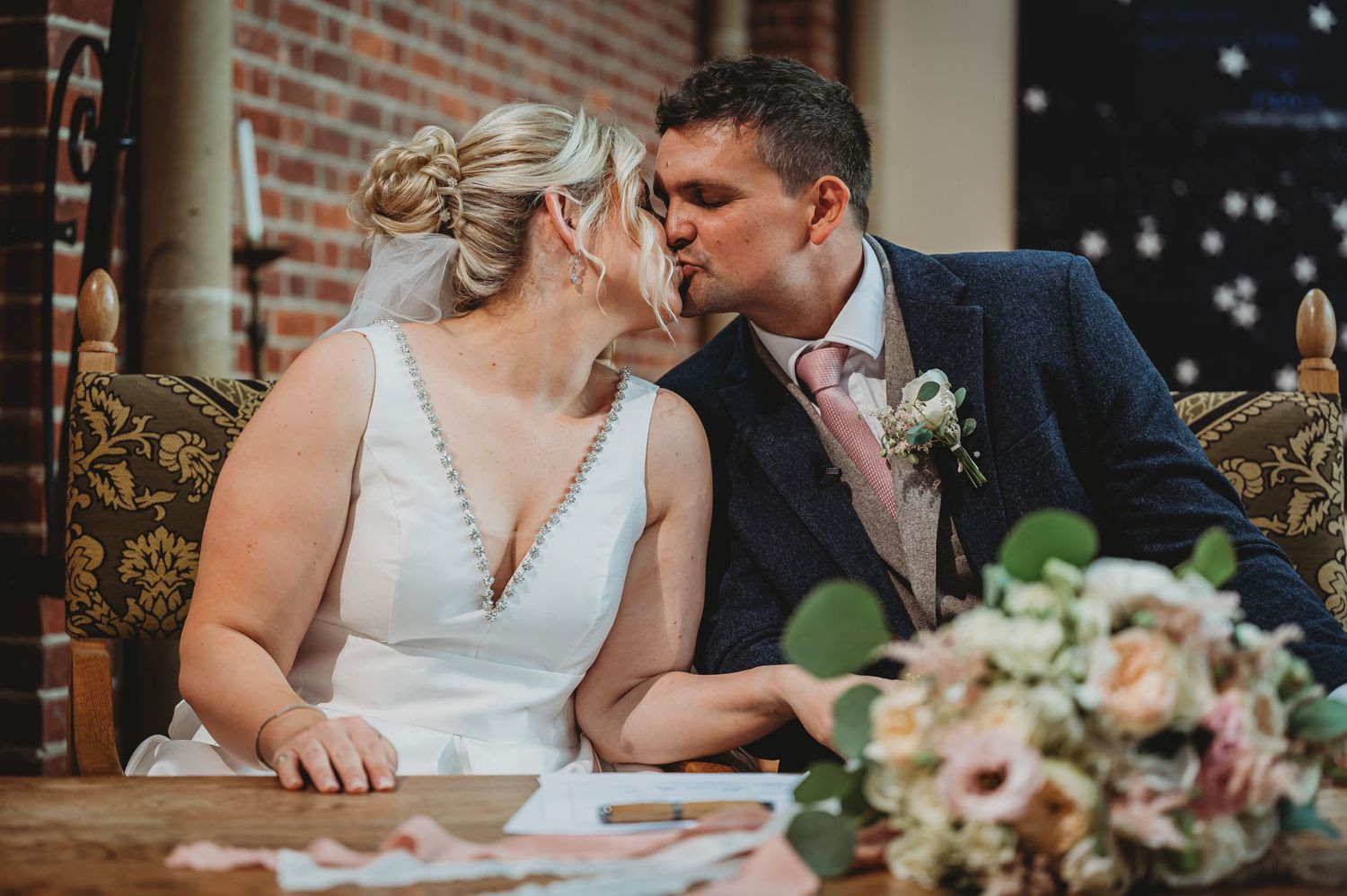 Newlywed couple shares intimate moment at rustic reception venue with brick walls and floral bouquet on table.