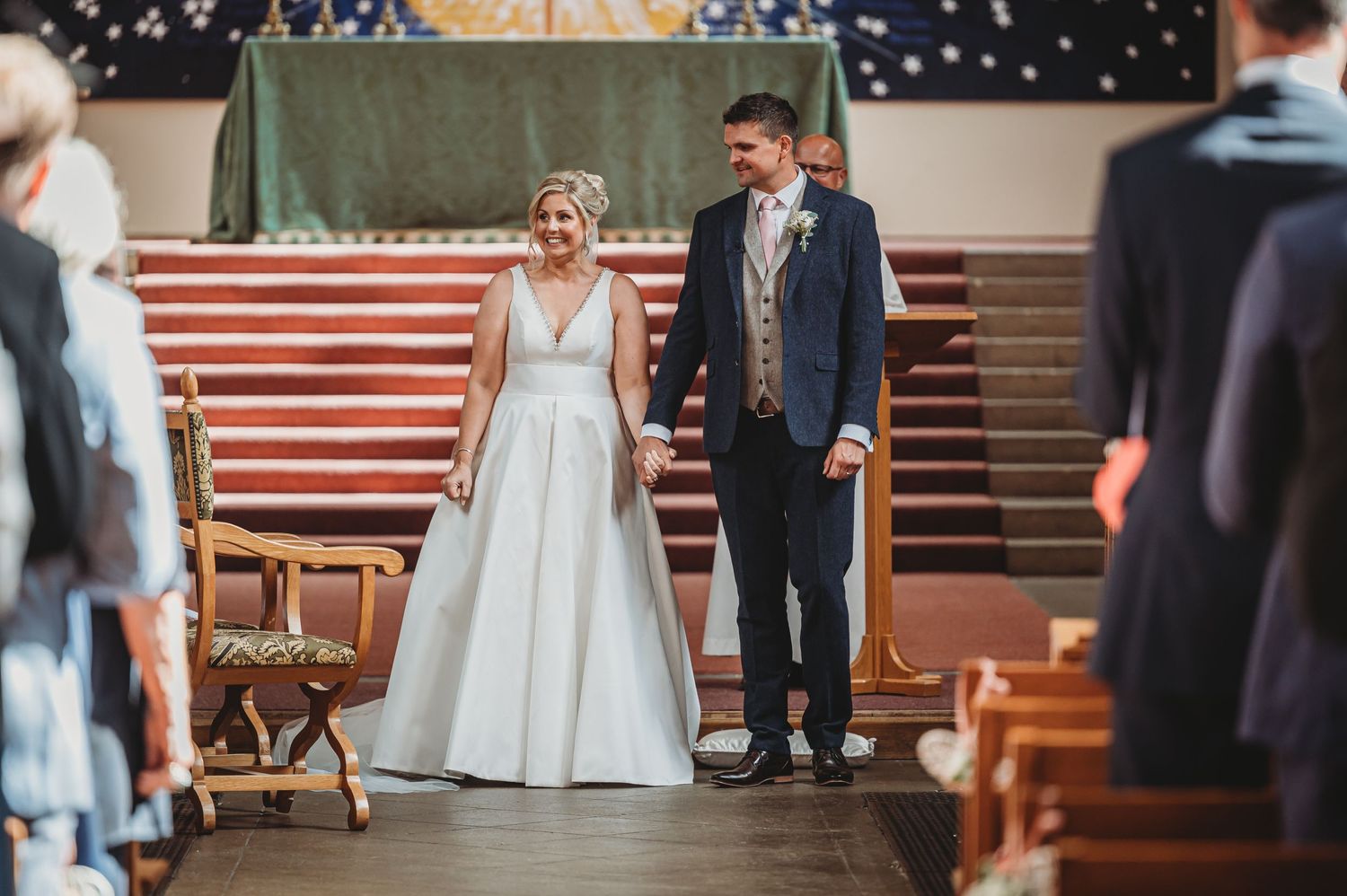 A couple stands together during their wedding ceremony in a church decorated with glowing lights and wooden pews.