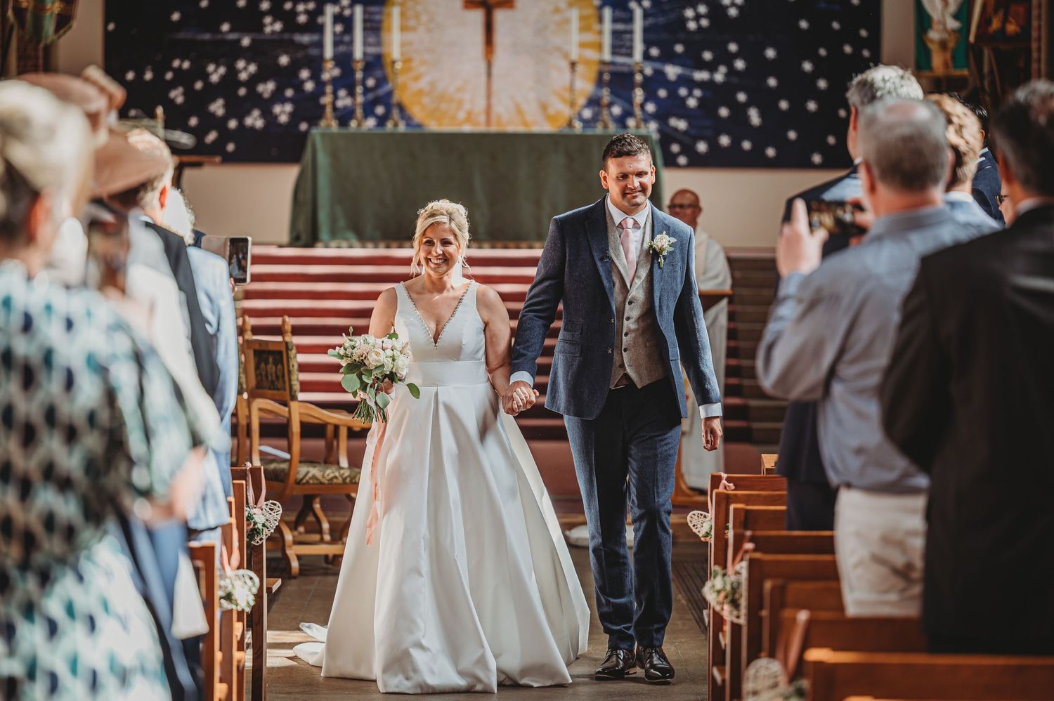 A couple walks down the church aisle during their winter wedding ceremony with twinkle lights in the background.