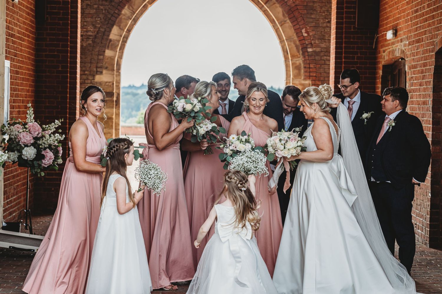 Wedding party gathers under brick archway with bridesmaids in pink dresses holding floral bouquets.