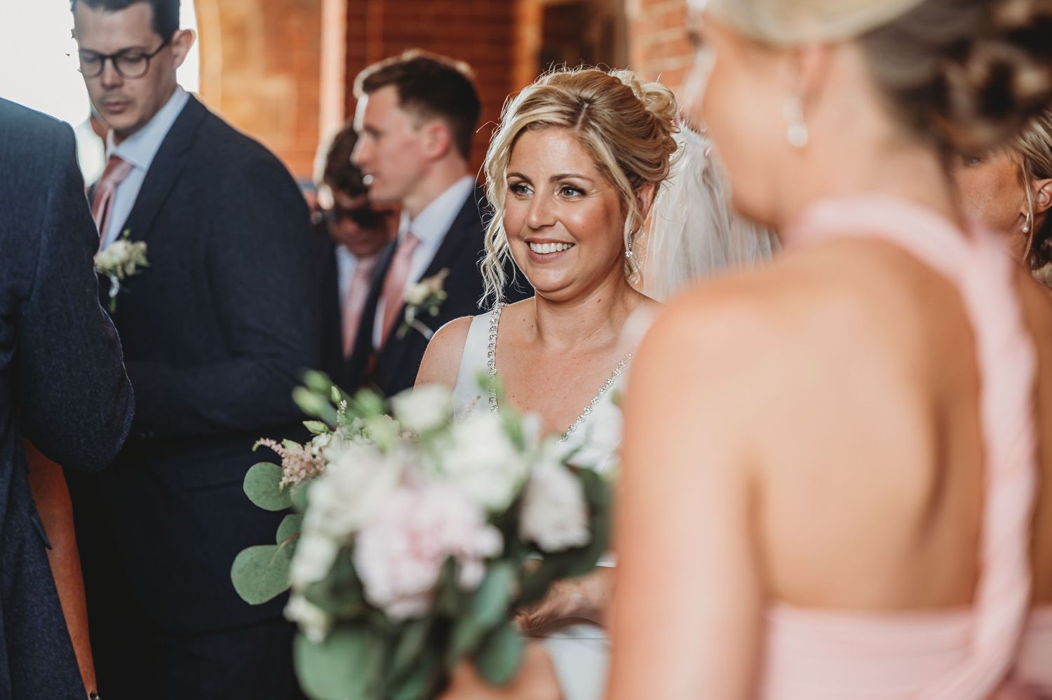Bride smiles joyfully while holding pink and white bouquet at indoor wedding ceremony.