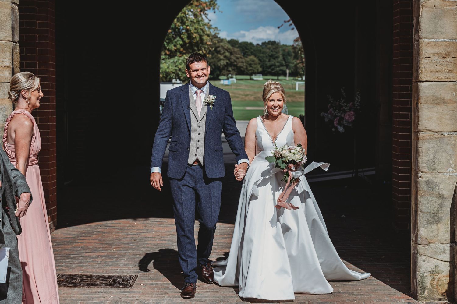 Newlyweds emerge from stone archway at outdoor venue with bride in white gown and groom in navy suit.