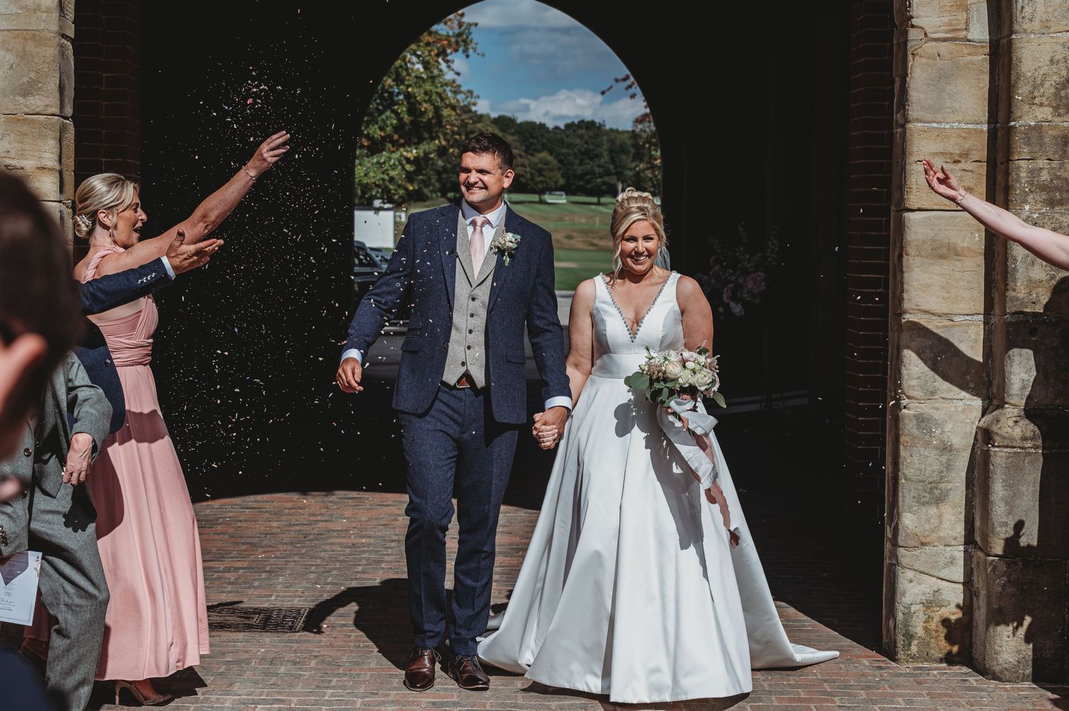 Wedding couple exits church door while guests throw confetti during sunny outdoor summer celebration.